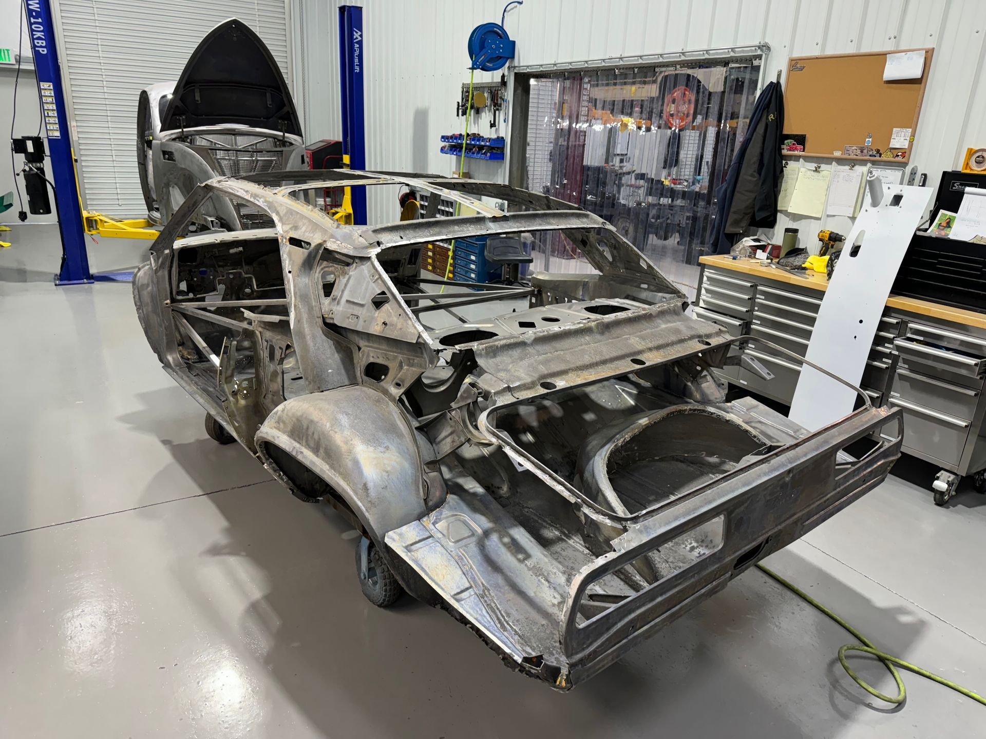 Stripped car body shell on a lift in an auto repair shop, with exposed frame and missing panels.