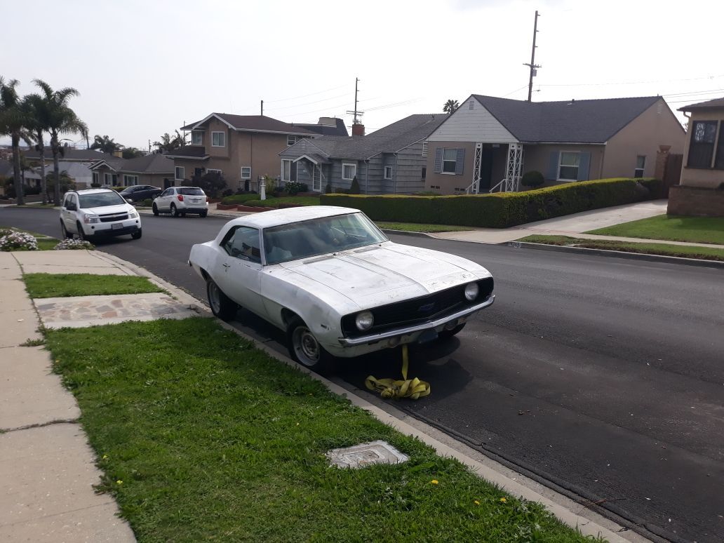 White car parked partly on a curb beside a suburban street, with a tow hook or wheel chock visible underneath.