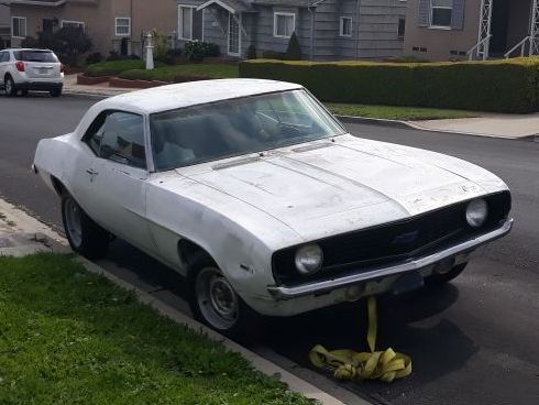White classic car parked at the curb with the front end lifted and a jack stand underneath.
