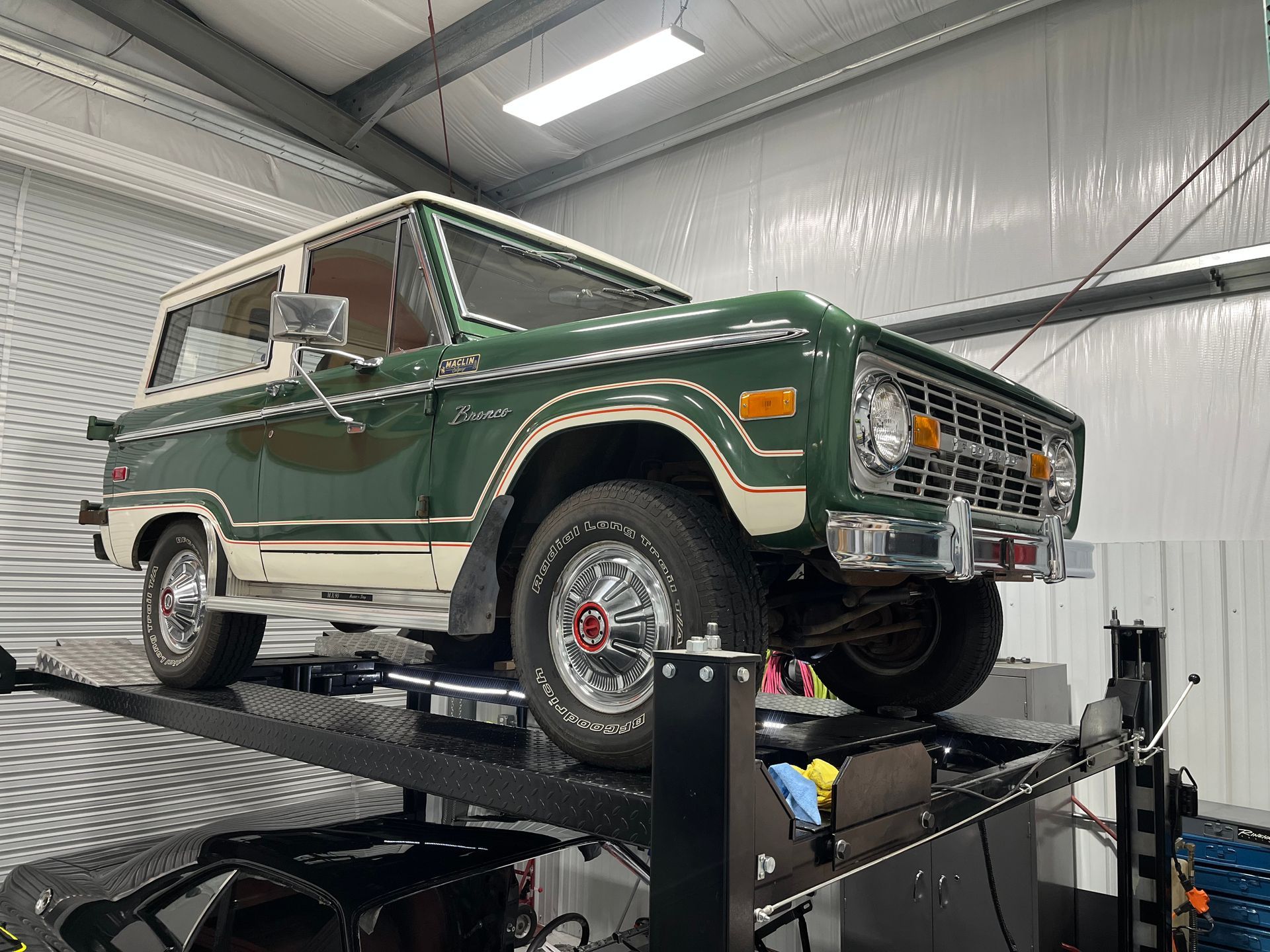 Classic green-and-white Ford Bronco on a lift in a garage, front wheel removed.