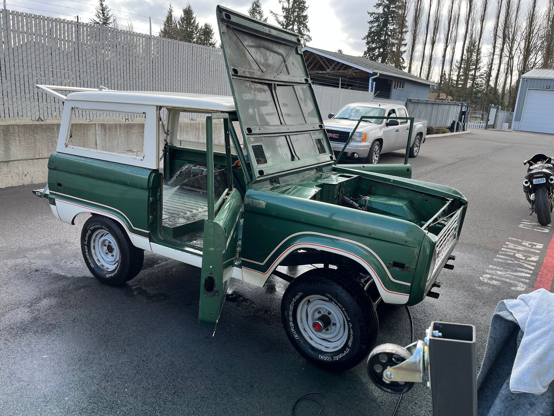 Green vintage off-road truck with hood open, parked in a lot near a building and another car.