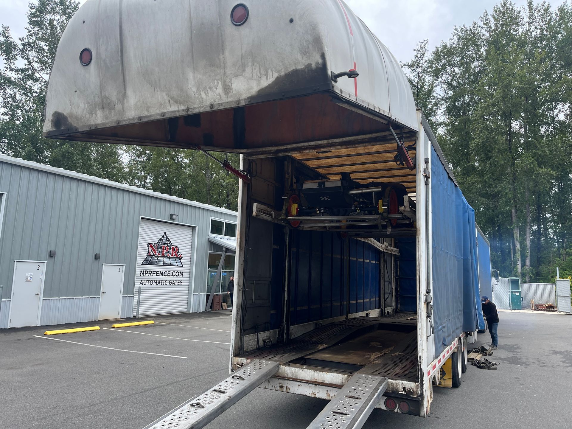 Open semi-trailer loading dock with raised roof, parked beside a gray warehouse; worker stands at right.