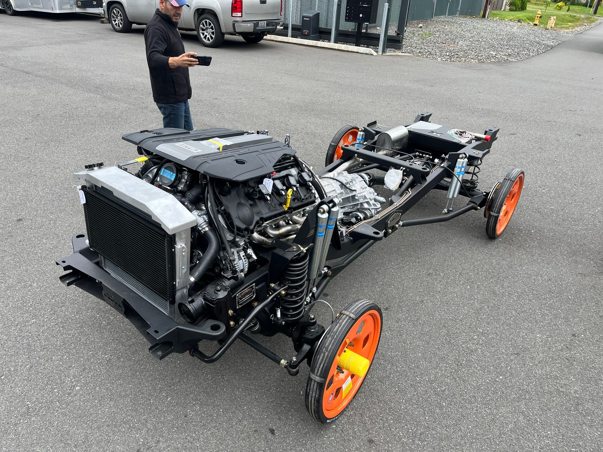 Stripped-down black car chassis with orange wheels parked on pavement, with a person standing nearby.