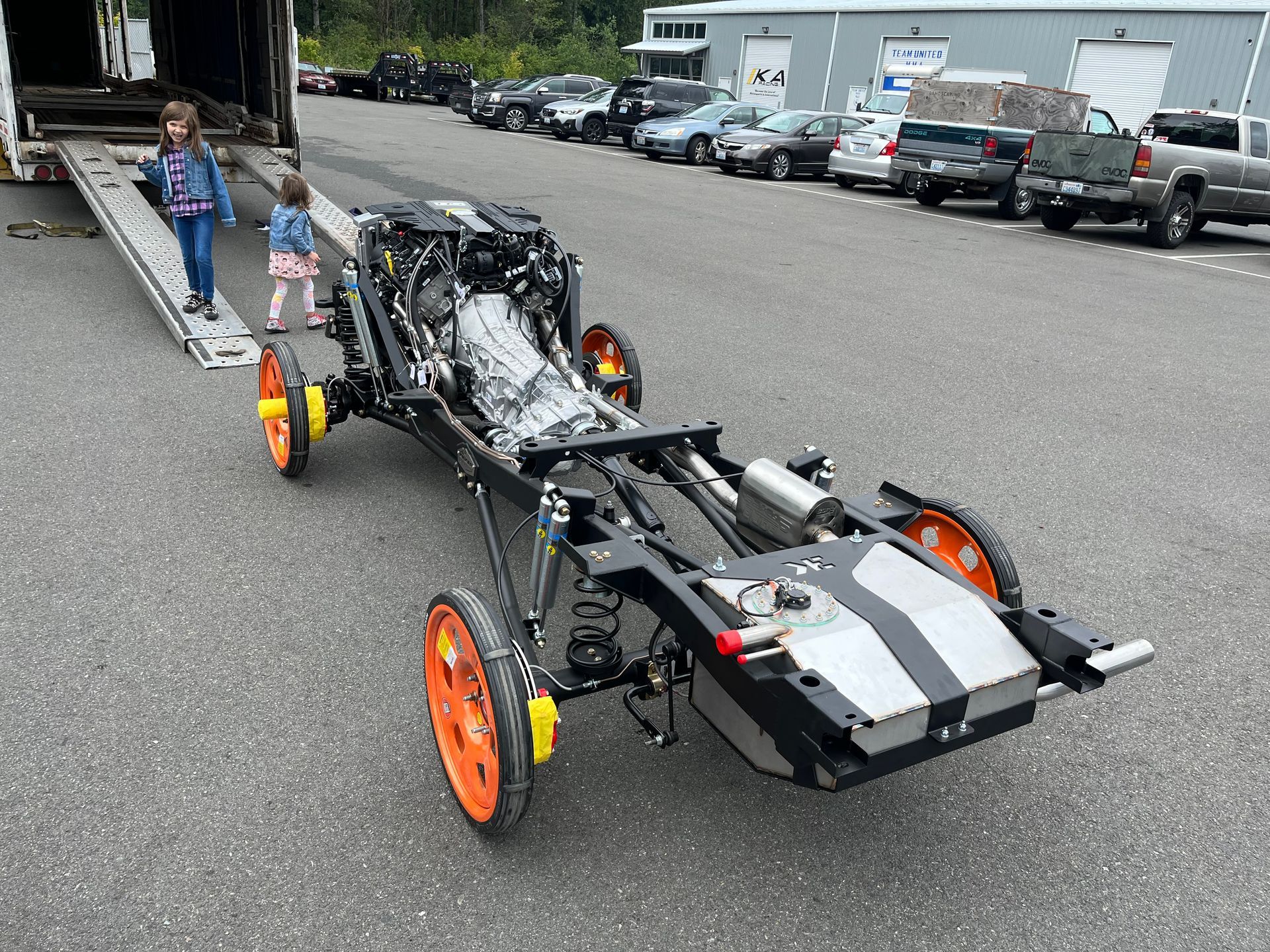 Small black-and-orange custom vehicle chassis on a trailer in a parking lot with two people nearby