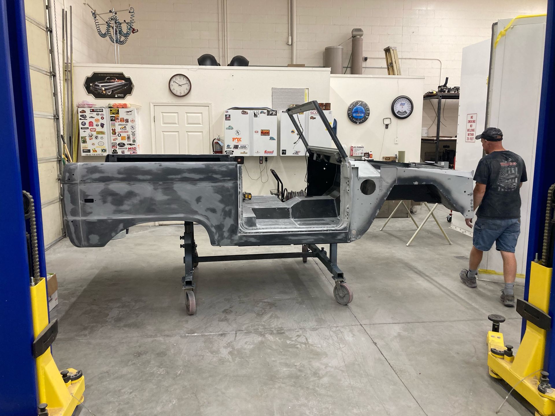 Partially stripped car body in a garage, with a person standing beside it and a lift in the foreground
