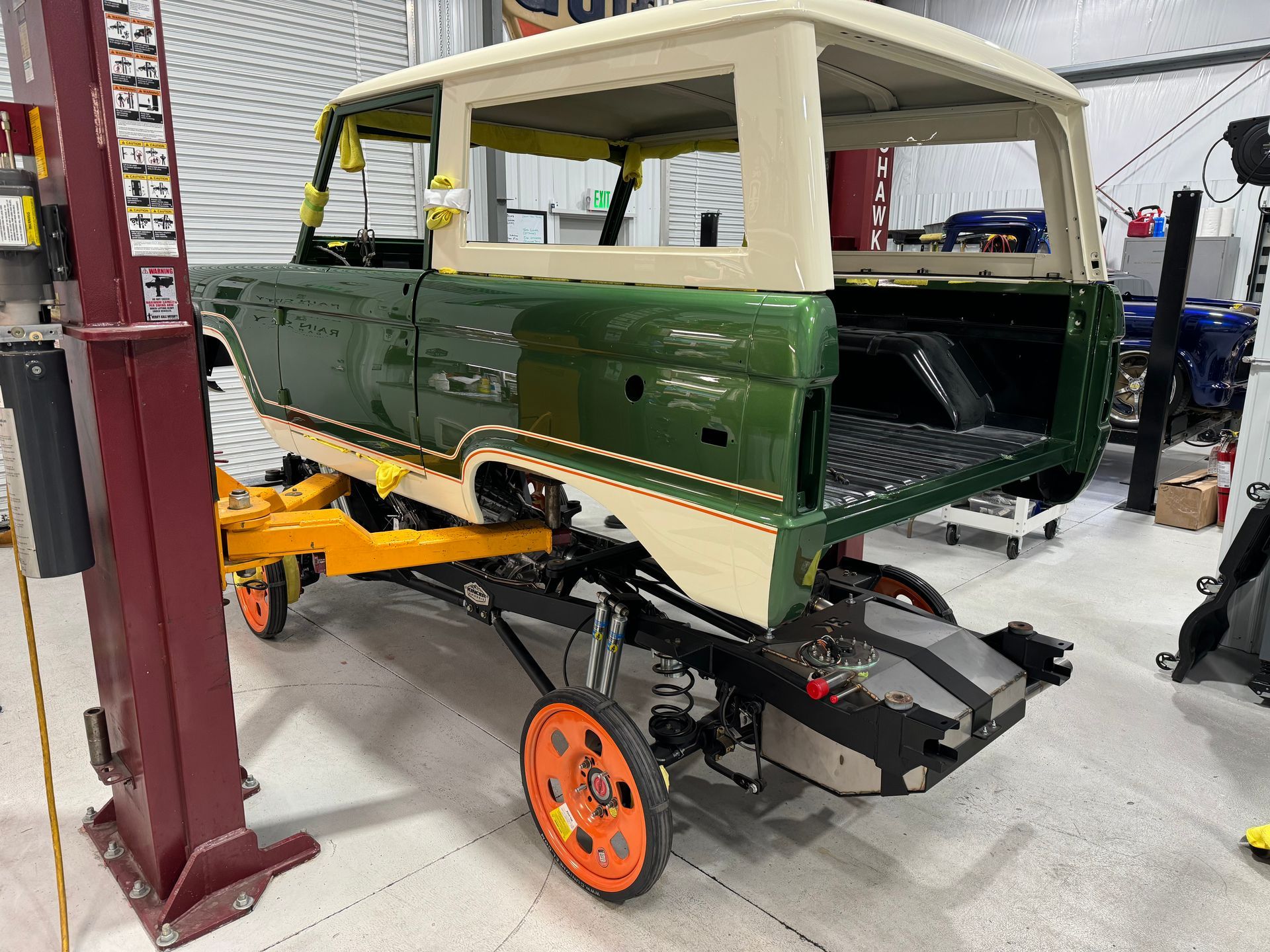 Vintage green vehicle body on a workshop lift, partially restored with exposed chassis and wheels.