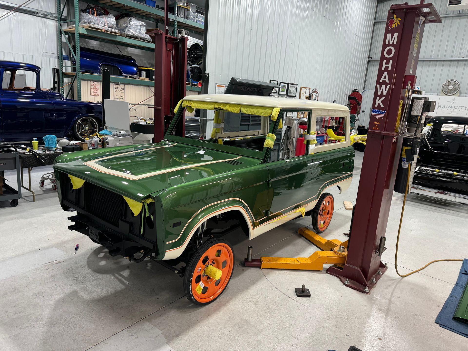 Green vintage SUV on a car lift in an auto shop, with orange wheels and exposed body panels.