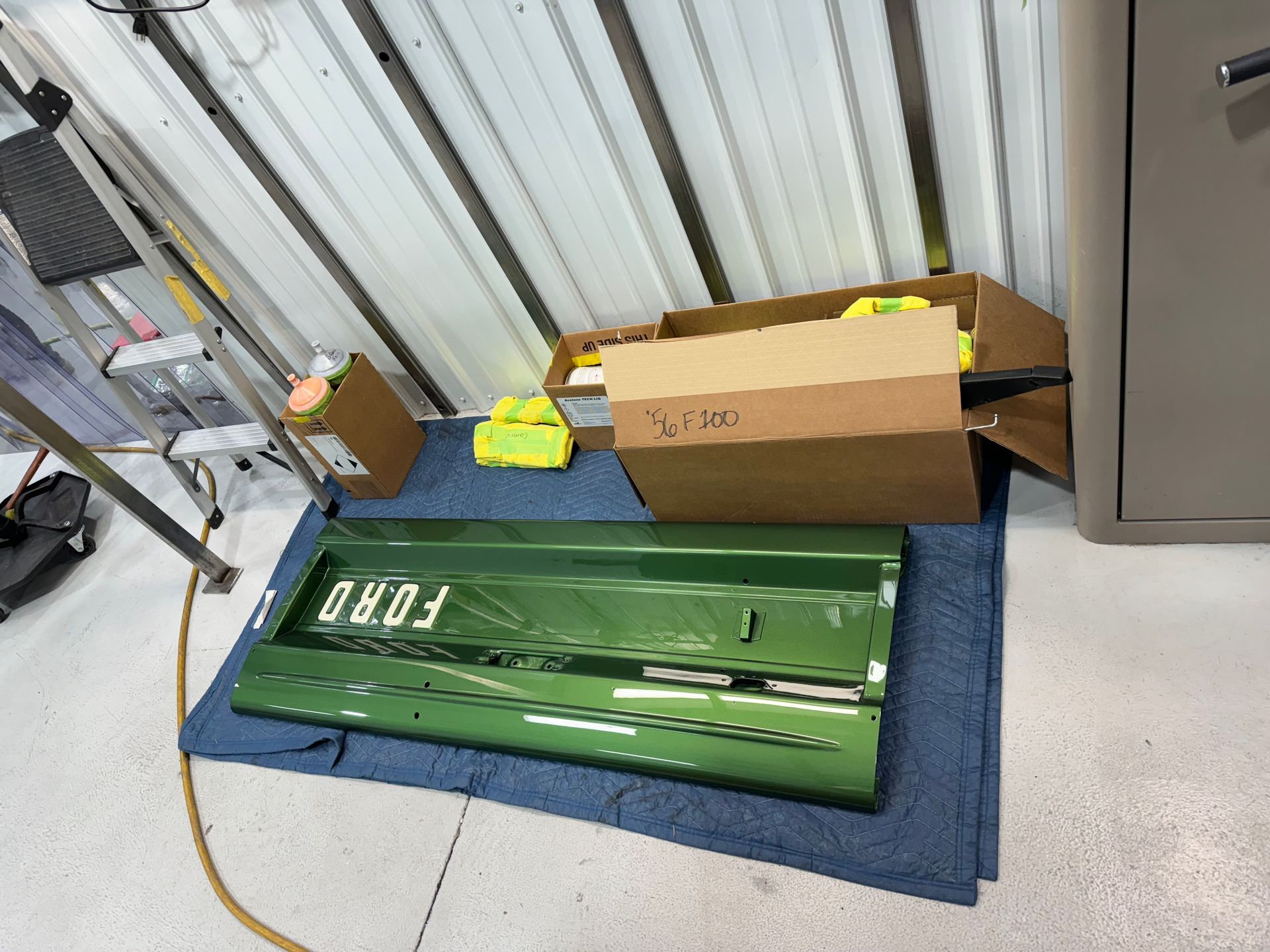 Green car hood on blue mat beside cardboard box and metal cabinet in a workshop