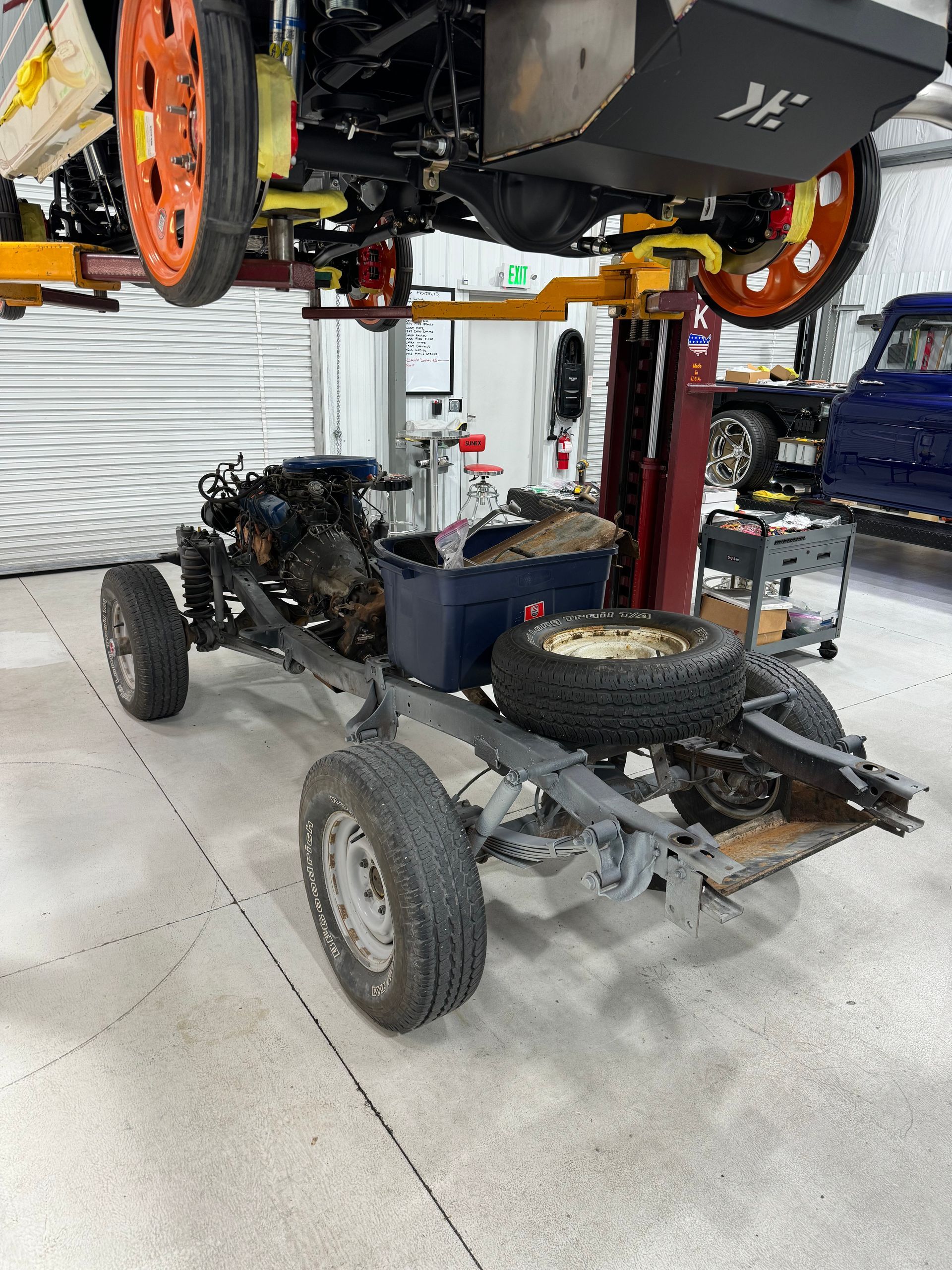 Vehicle chassis on a workshop lift with exposed frame, wheels, and tools in a garage.
