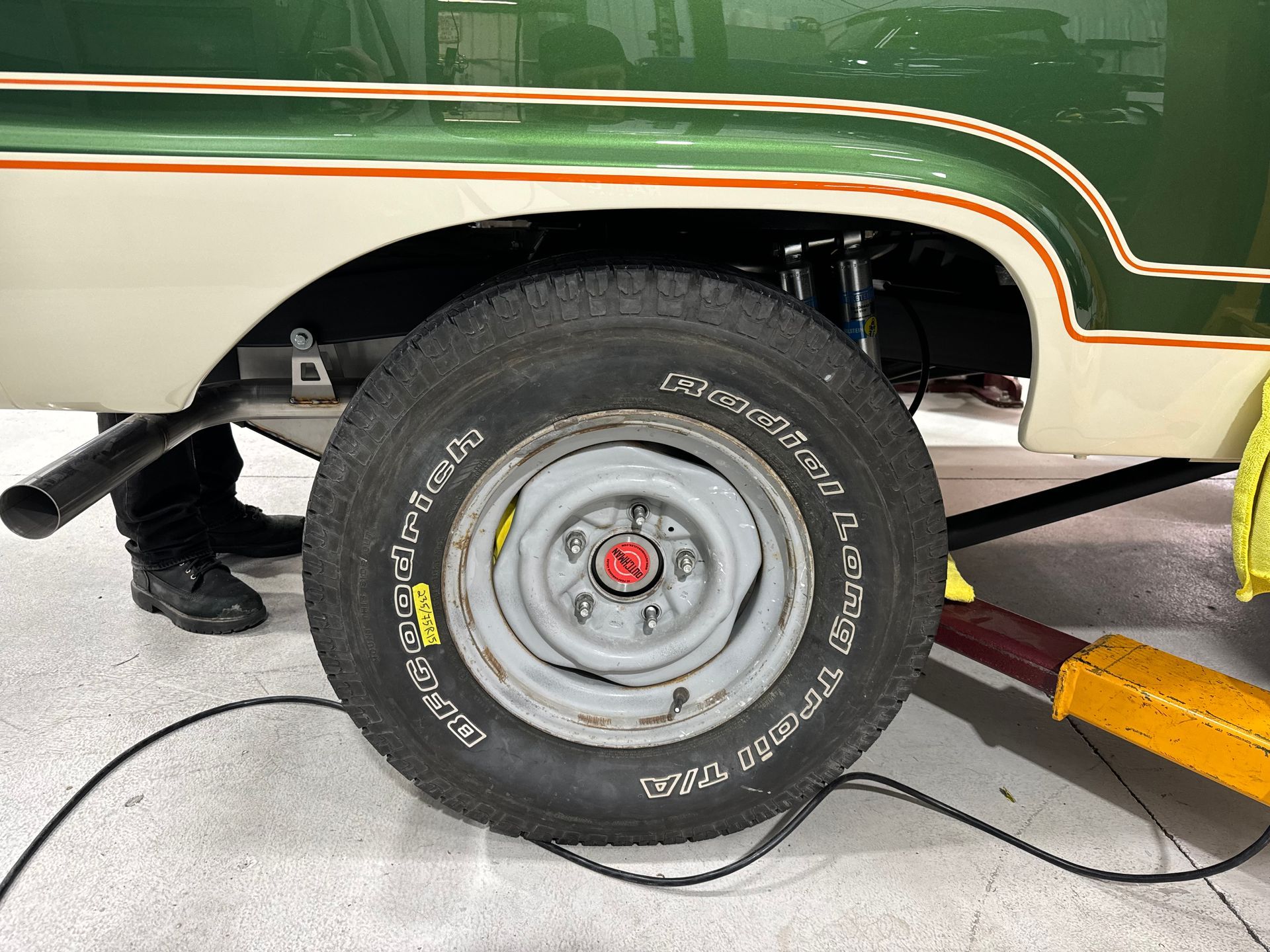 Close-up of a vehicle's rear wheel on a lift, with a jack stand and mechanic's leg visible.