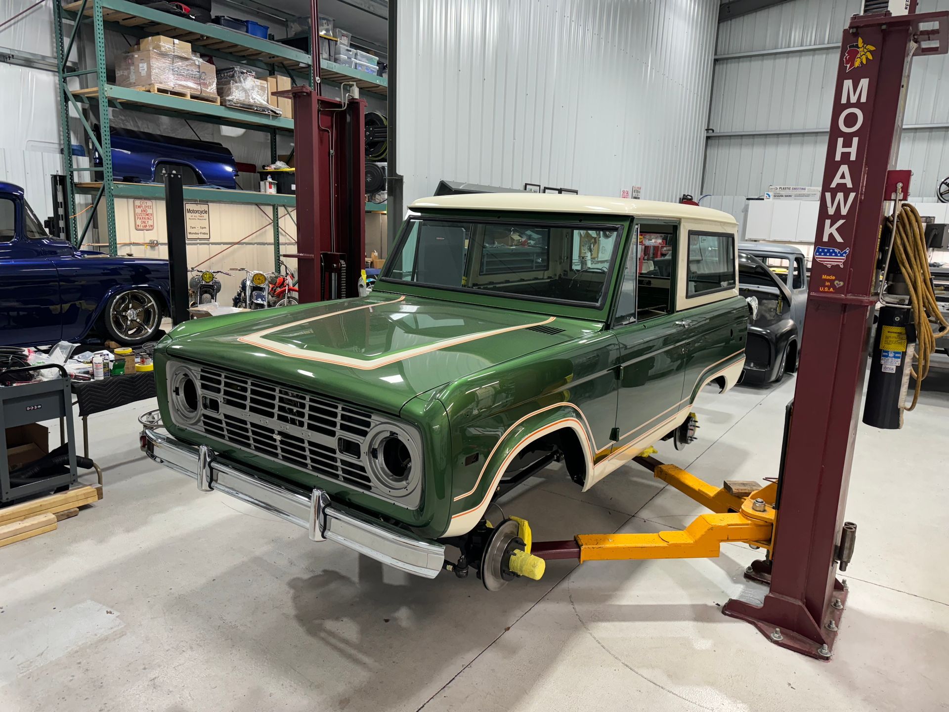 Green classic Bronco on a lift in an auto shop, with a red Mohawk post and blue car nearby.