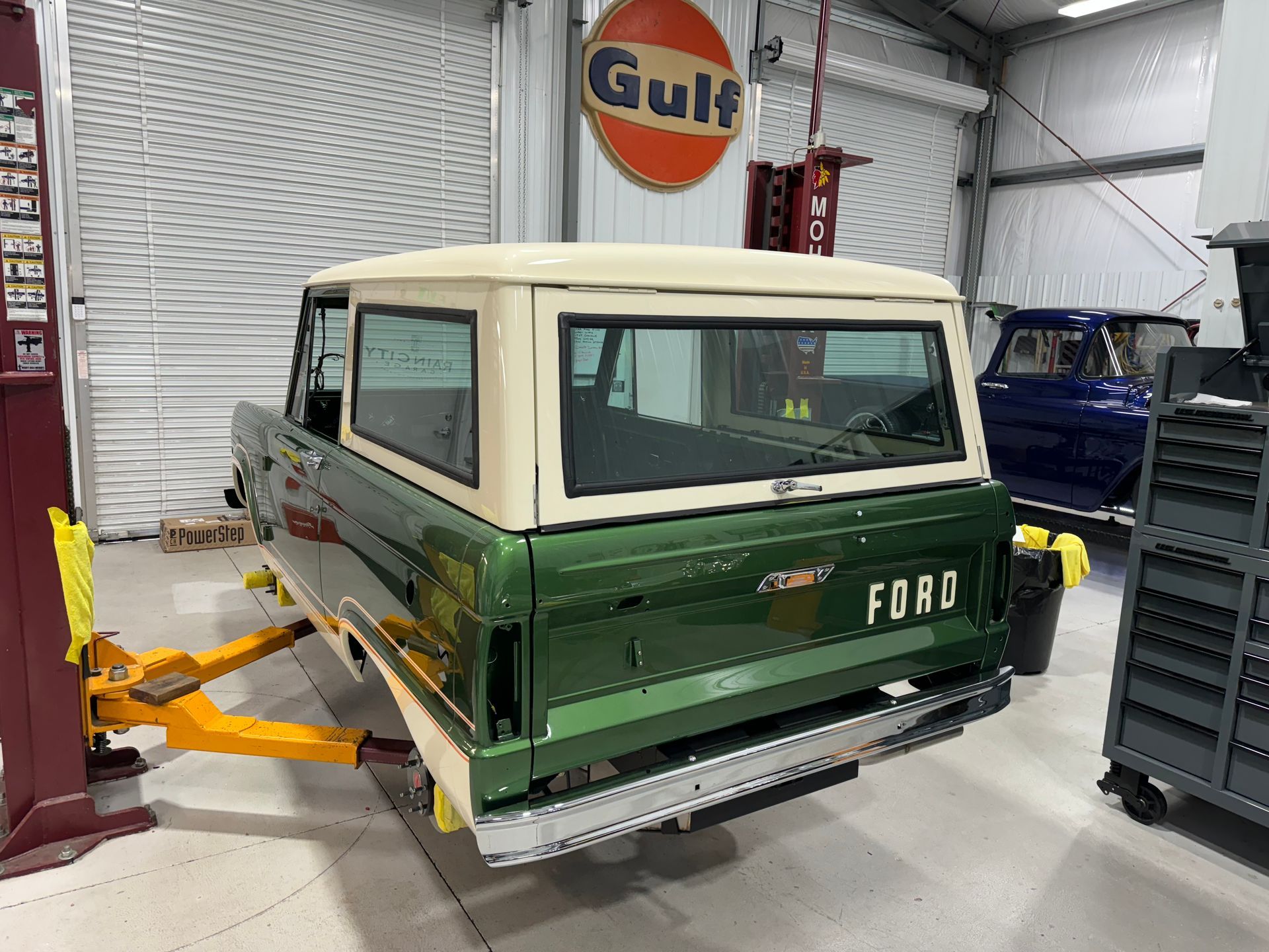 Green and white Ford Bronco on a lift in an auto shop, rear view.