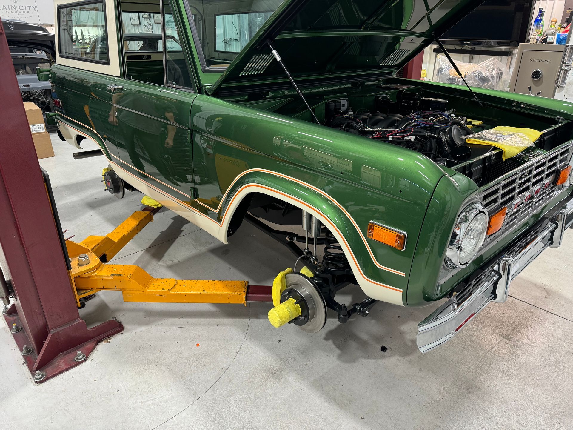 Green classic car on a lift in a garage, front wheel removed and hood open during repair.
