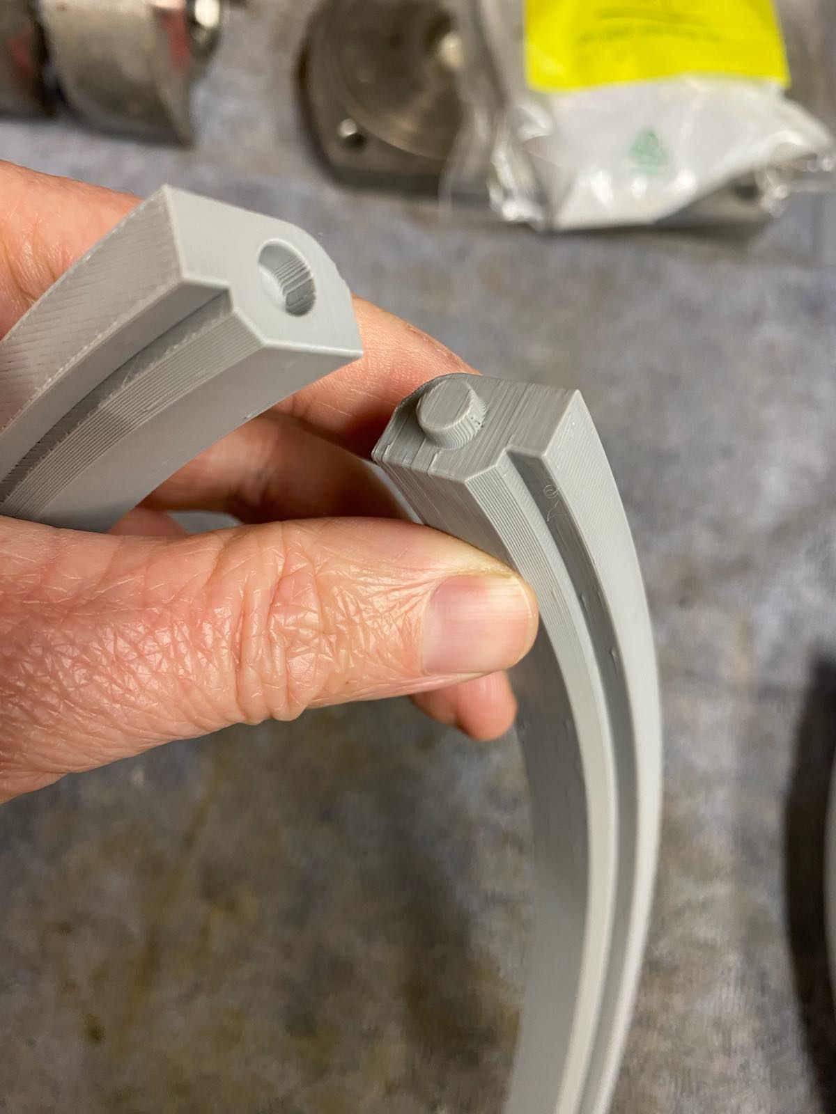Hand holding two gray 3D-printed curved parts with holes, in a workshop setting