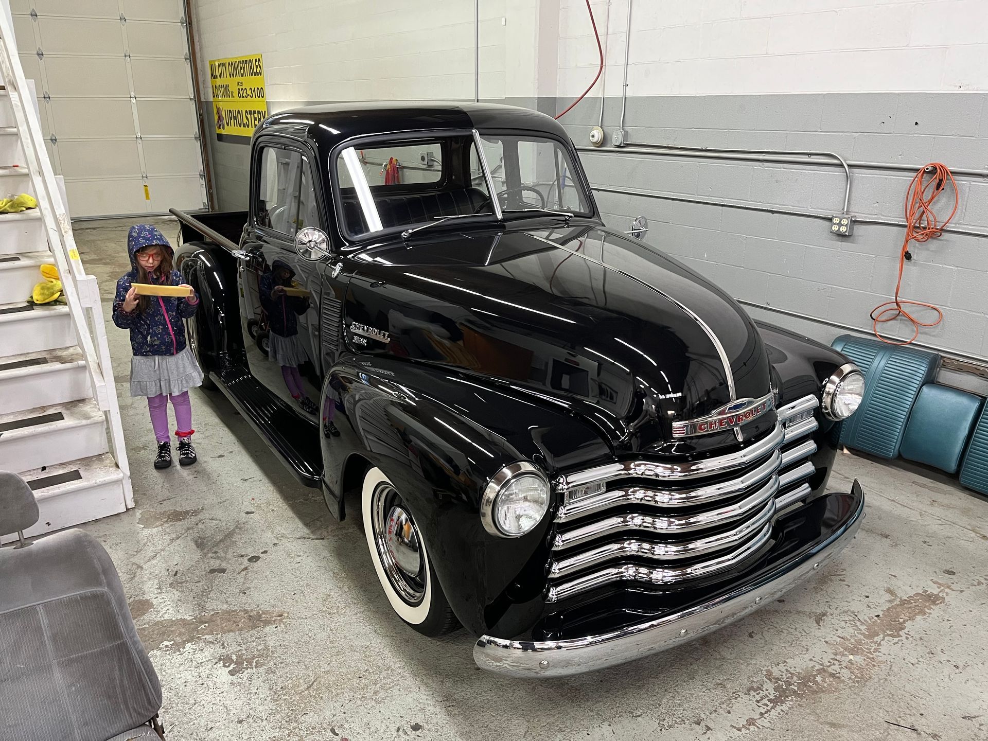 Black vintage pickup truck parked in a garage beside a child and a blue couch.