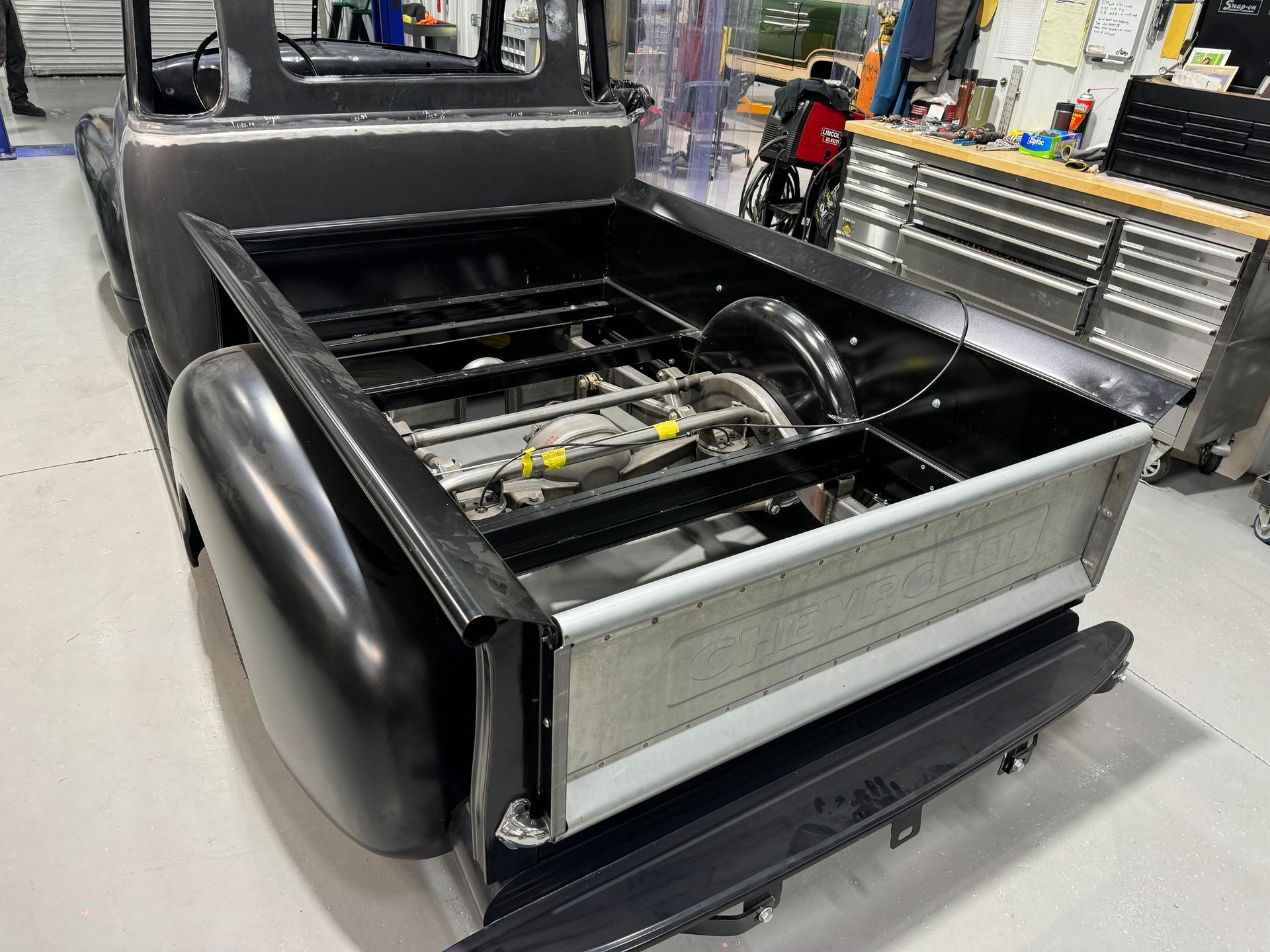 Black upholstered vehicle seat assembly in a workshop, with exposed metal frame and machinery visible behind it
