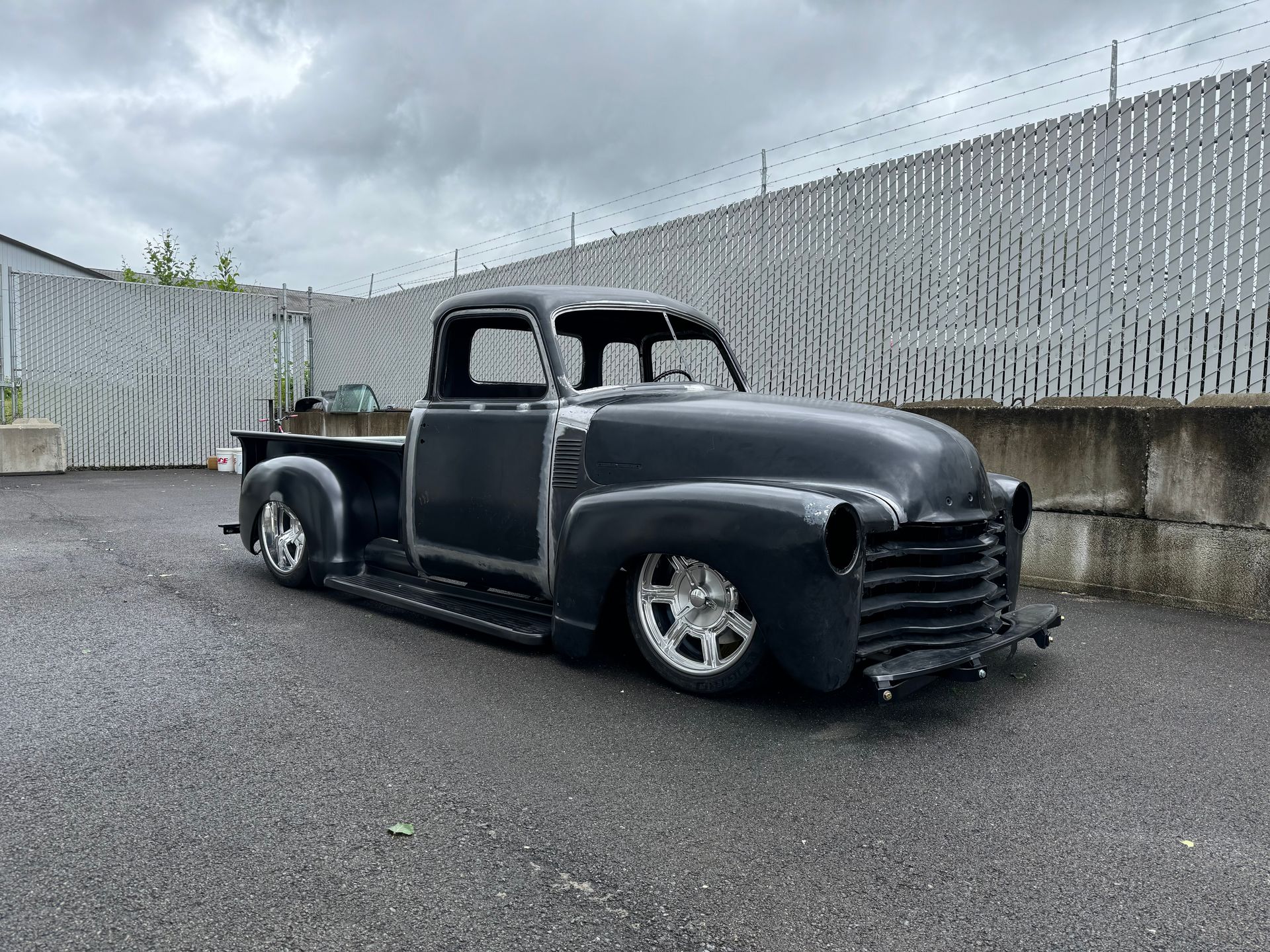 Black lowered vintage pickup truck parked on wet pavement by a tall fence under cloudy skies