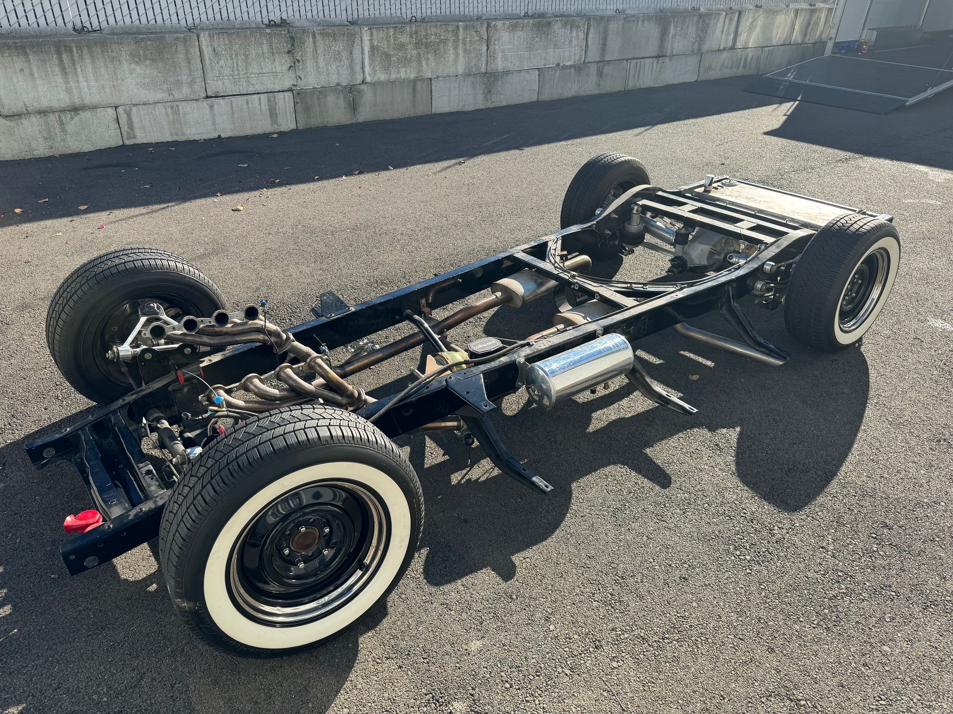 Bare electric vehicle chassis on asphalt, viewed from the rear-left side, with exposed frame and wheels