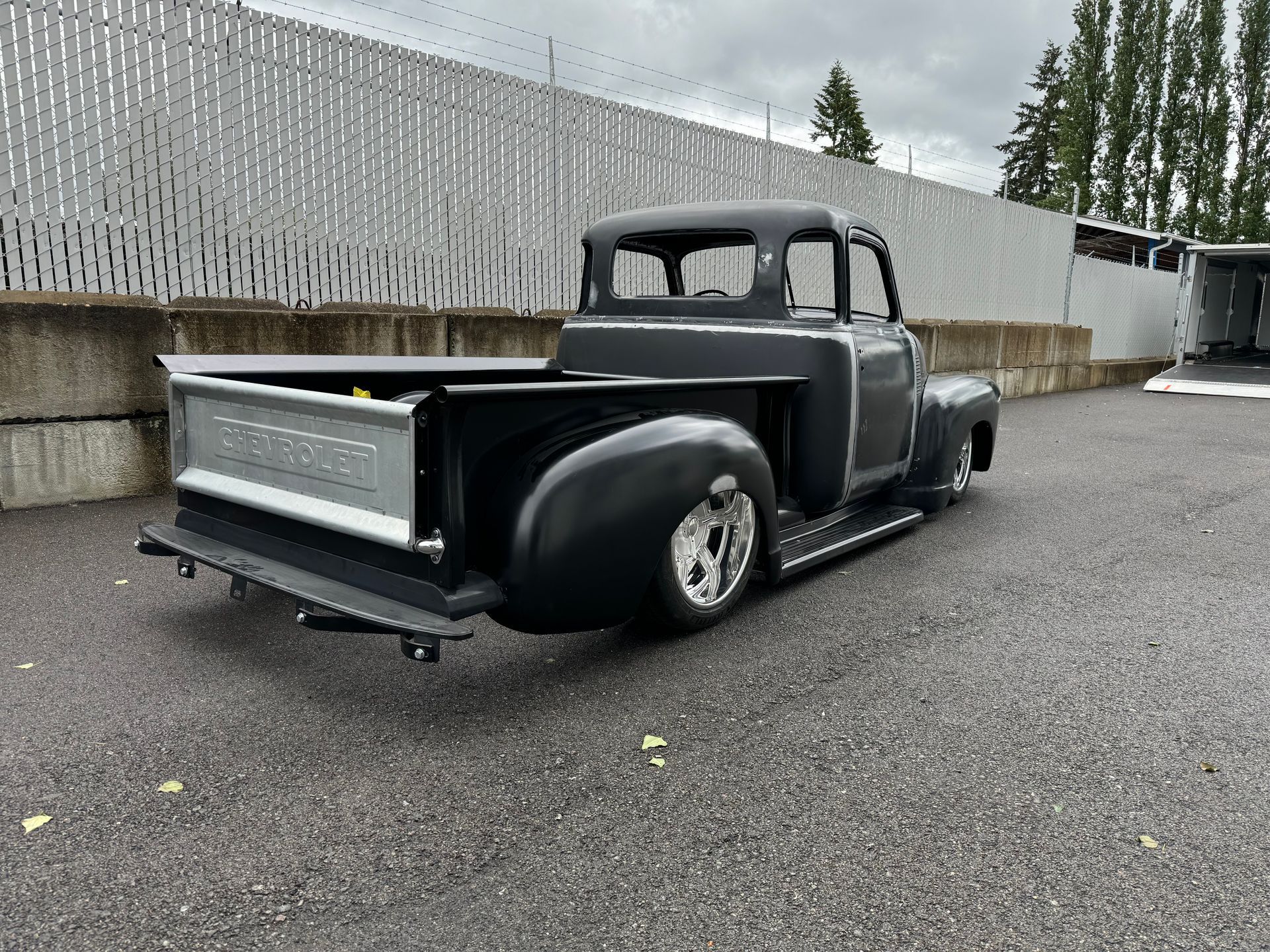 Custom gray lowrider pickup truck parked on wet pavement beside a concrete wall and fence.
