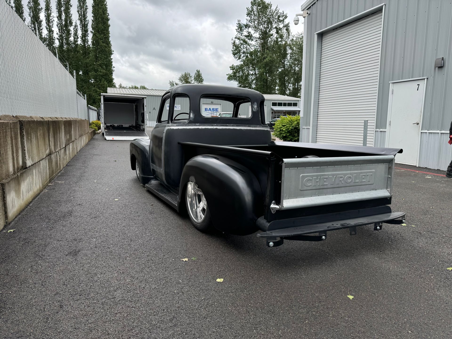 Black vintage pickup truck parked on a wet driveway beside a gray building, with another truck in the background