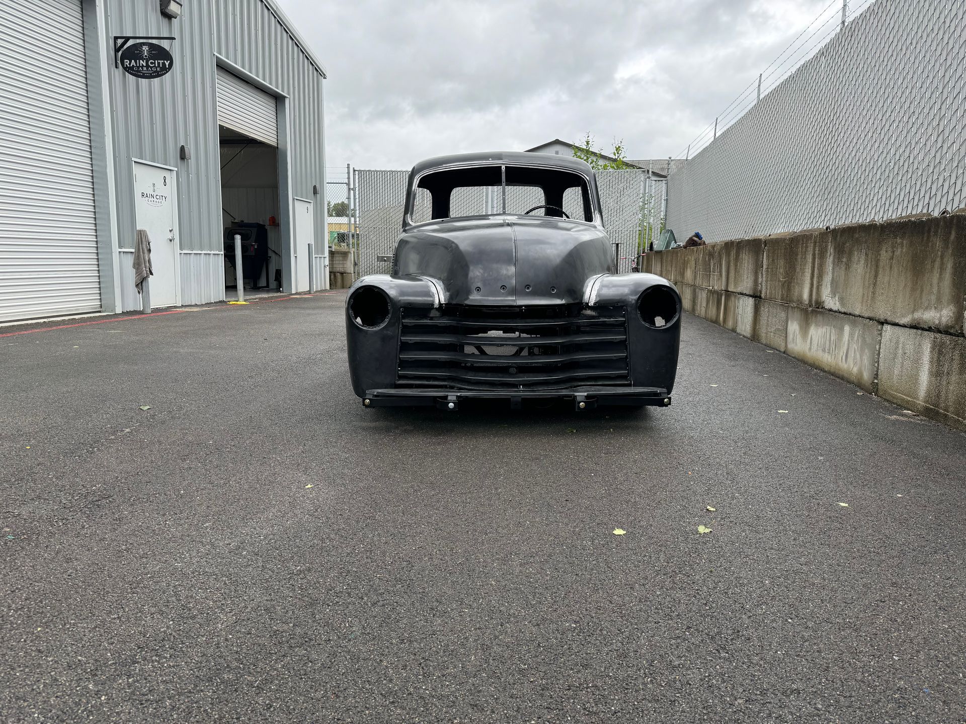 Black vintage truck parked on an industrial road between metal buildings under cloudy skies