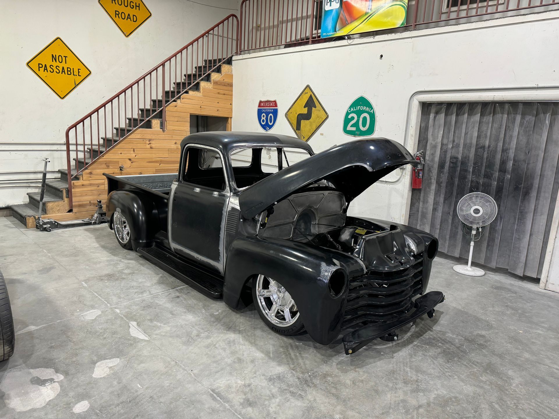 Black vintage pickup truck with hood open in a garage, with a staircase and road signs behind it