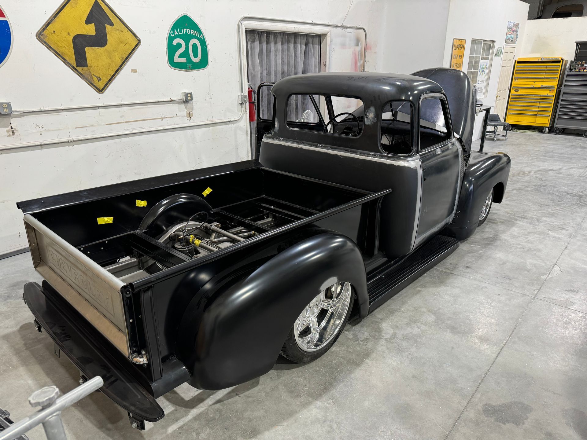 Black vintage pickup truck in a garage, rear bed open with a spare tire visible.