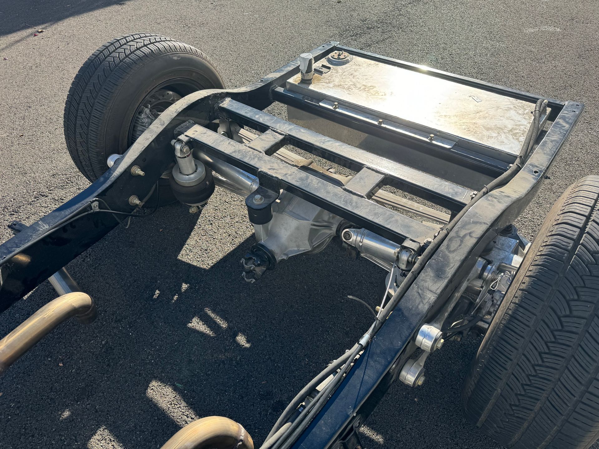Chassis frame of a vehicle on asphalt, showing rear wheels and suspension components.