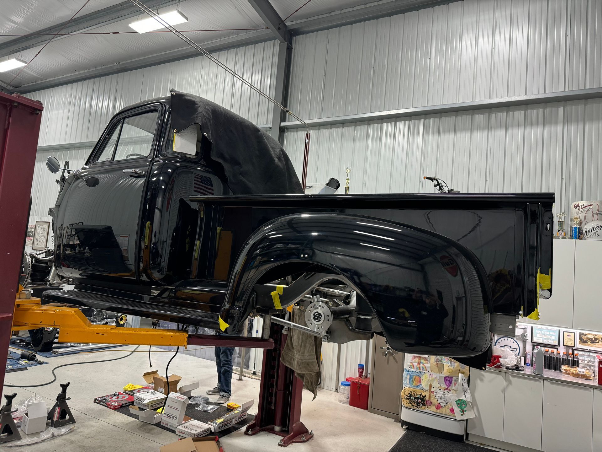 Black pickup truck body on a lift inside an auto repair shop, with wheels removed and exposed suspension.