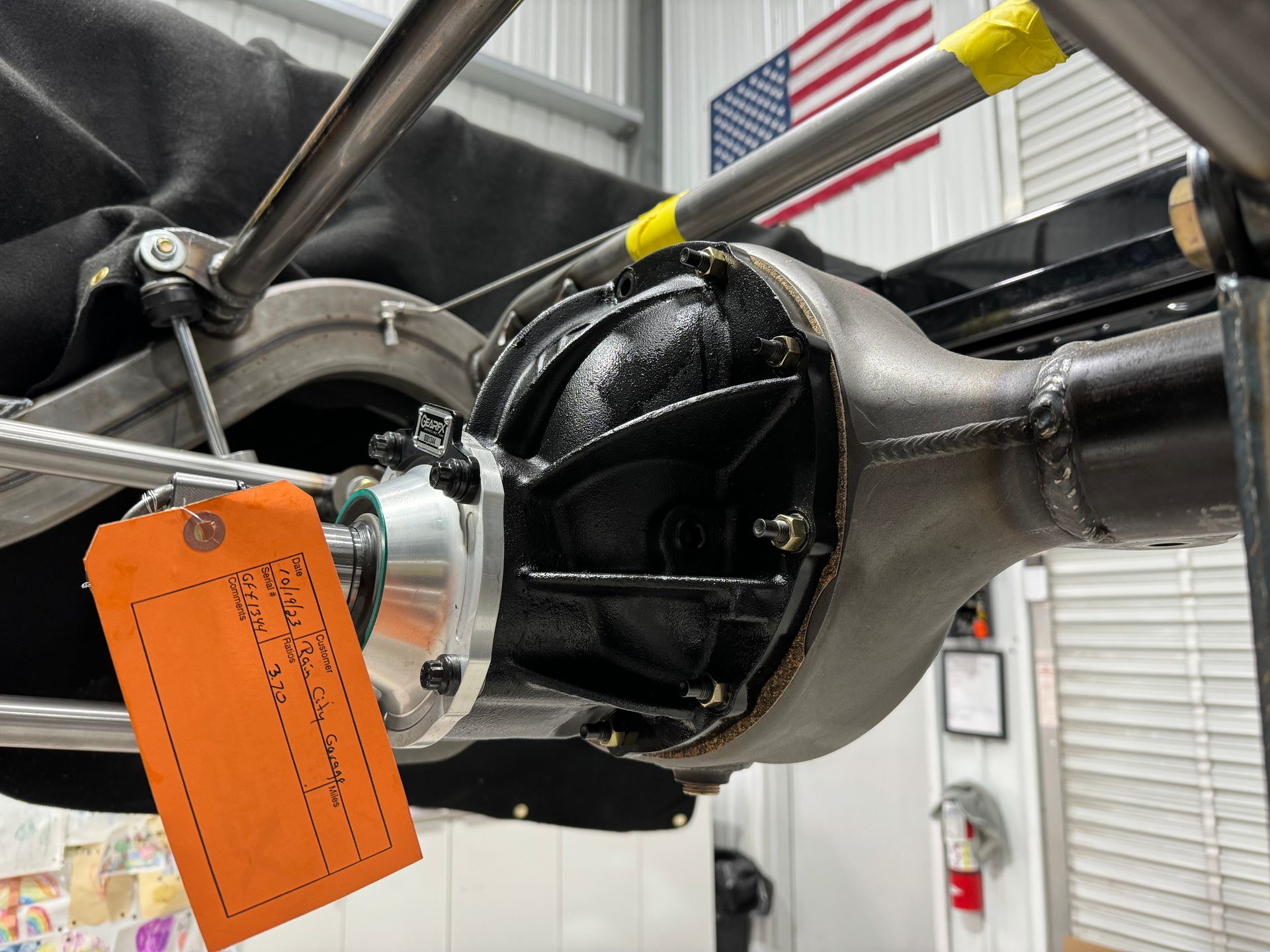 Close-up of a vehicle axle assembly with a bright orange inspection tag attached in a workshop.