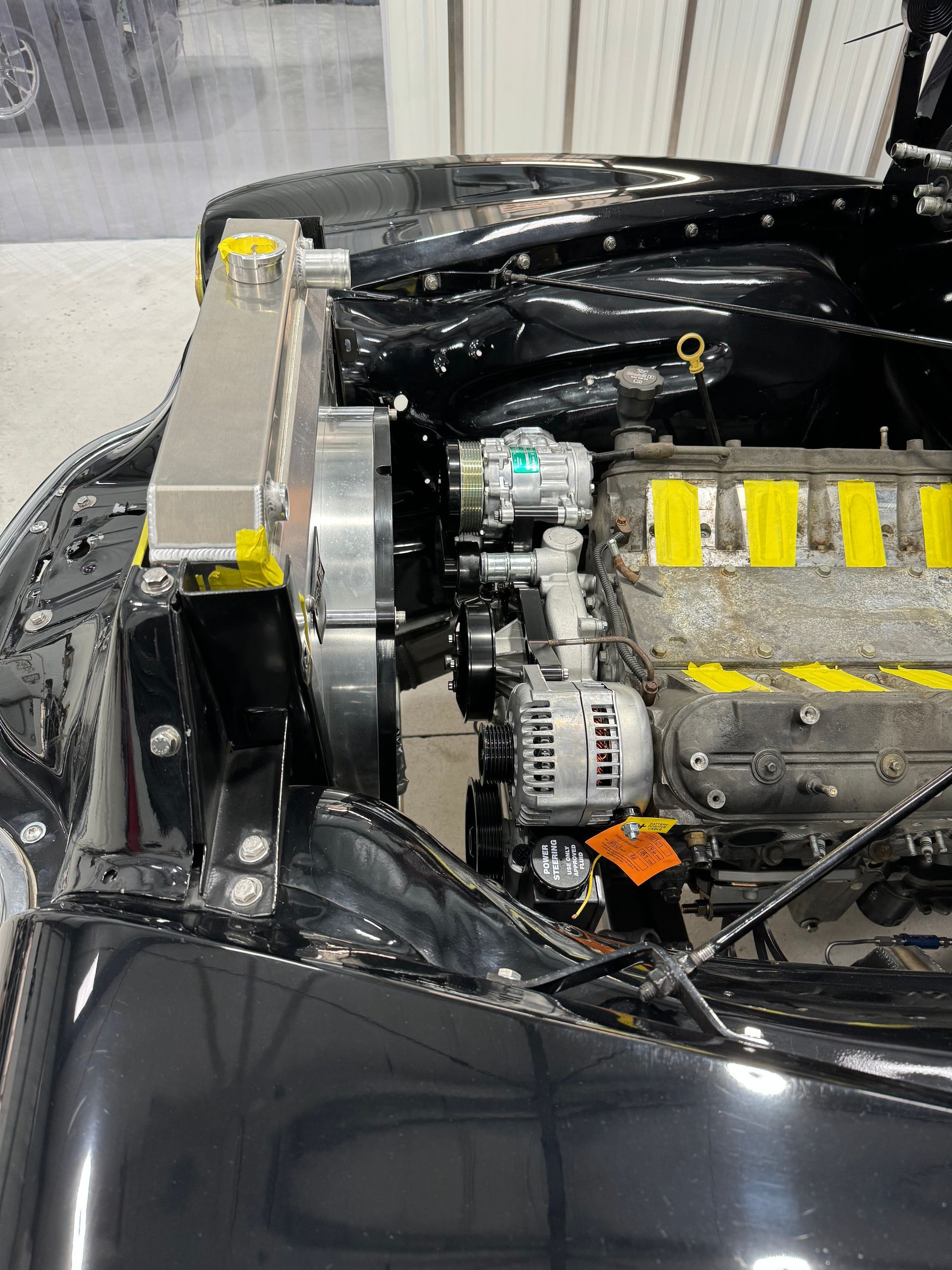 Open car engine bay with exposed silver engine, black body panels, and yellow tape markers