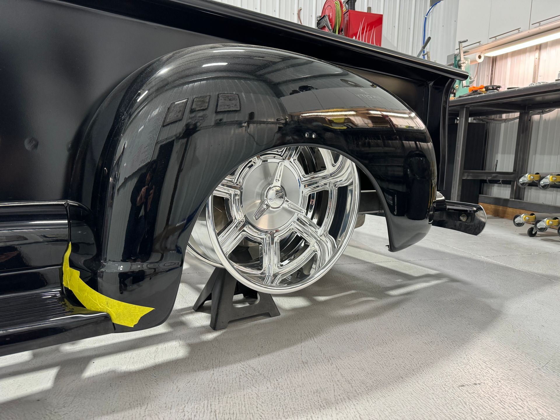 Close-up of a glossy black custom truck bed with chrome wheel and yellow brake caliper in a garage