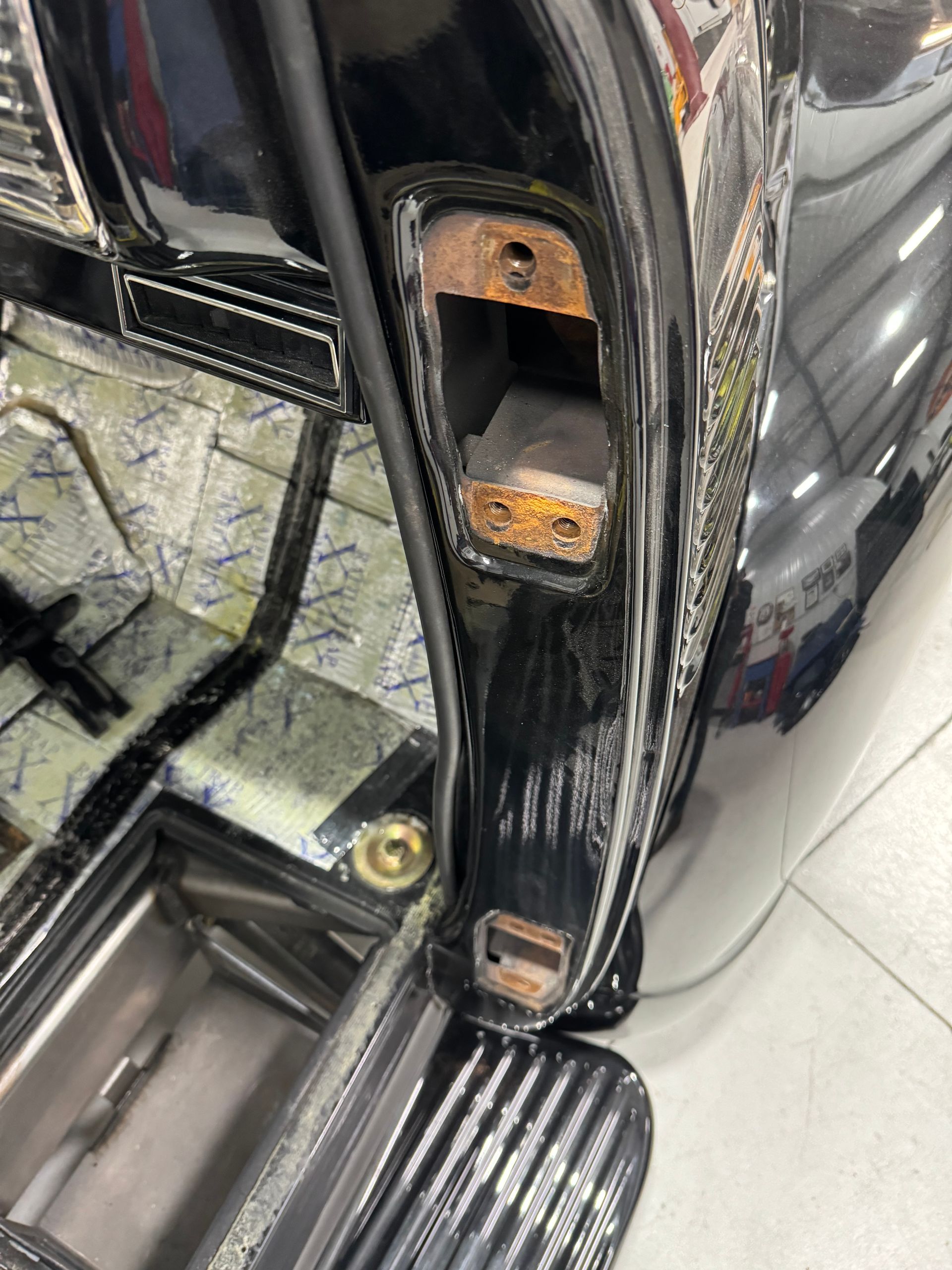 Close-up of a black car door hinge with rusted bolts and scuffed paint near the wheel well