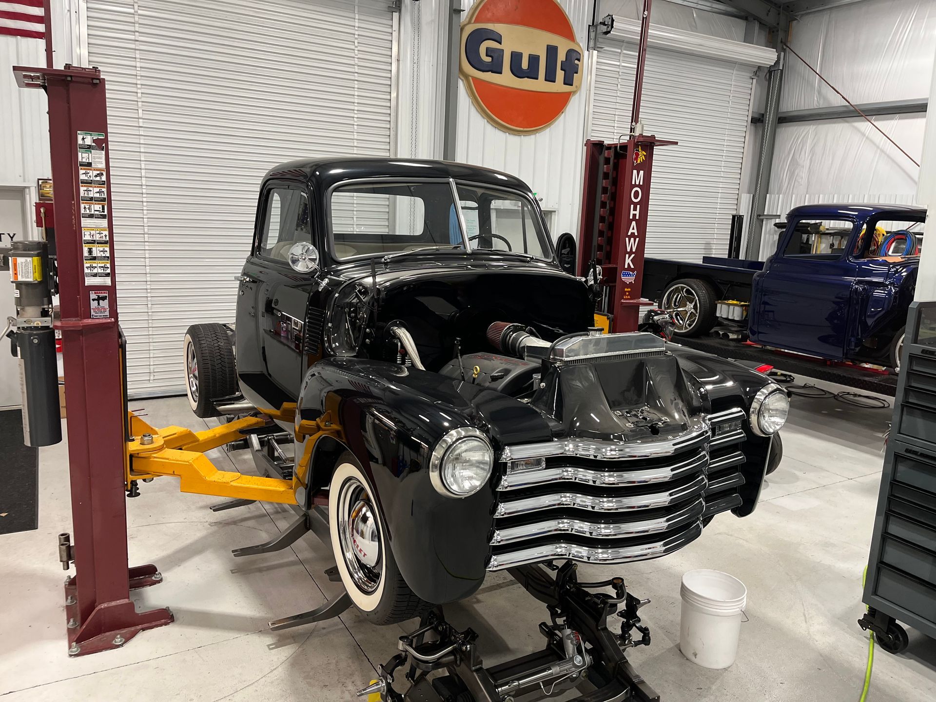 Black vintage pickup truck on a lift in a garage, with another classic truck in the background.