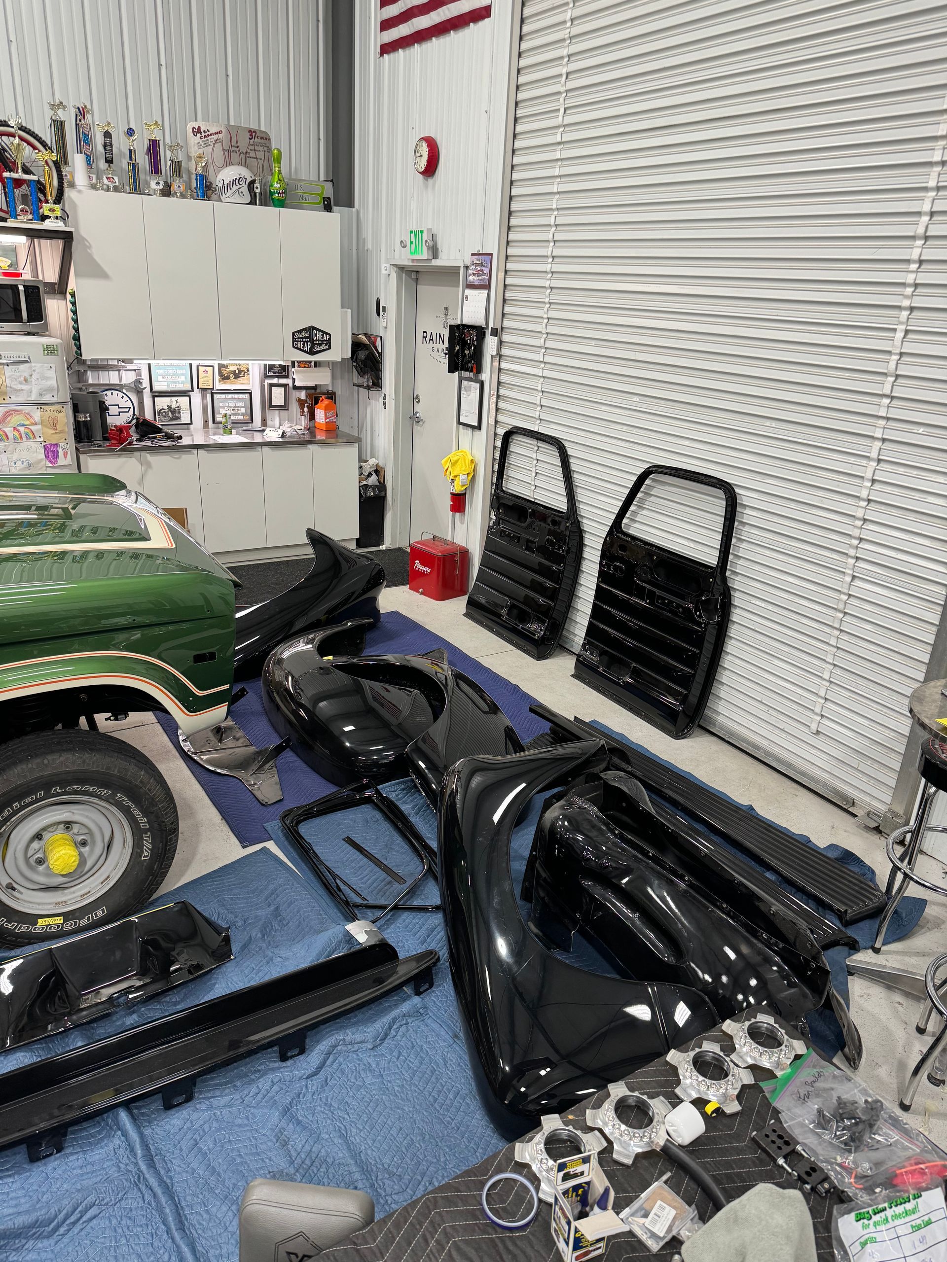 Garage/workshop cluttered with car parts, tools, and a green vehicle against a white wall