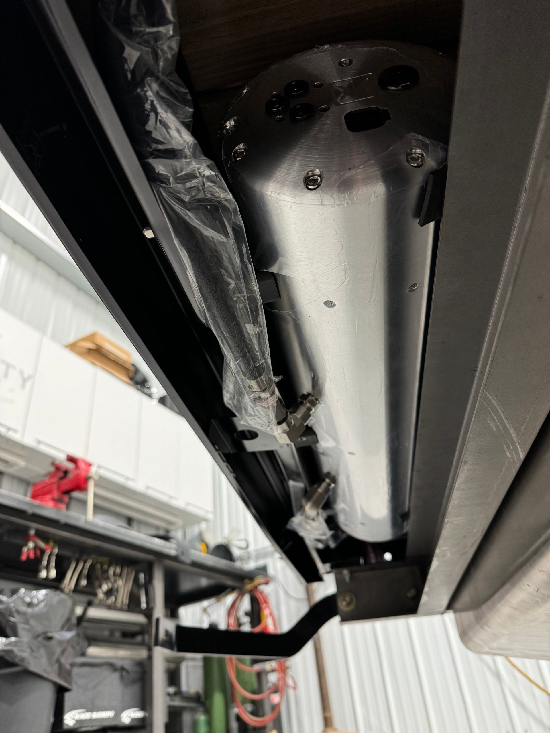 Underneath a vehicle, a silver exhaust or muffler assembly is visible on a lift.