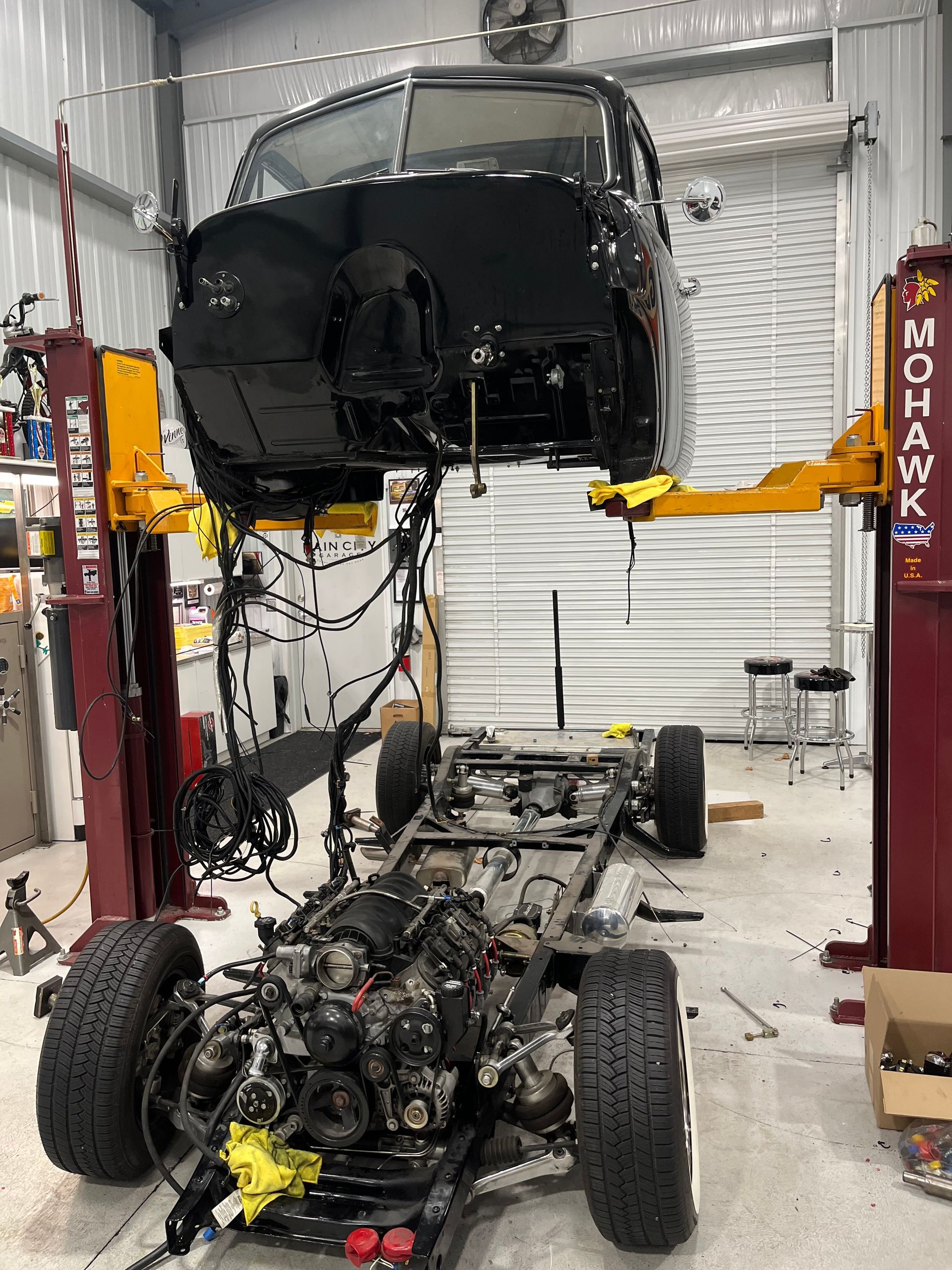 Car lifted on a hoist above a stripped chassis in an auto repair shop, with engine and suspension exposed.
