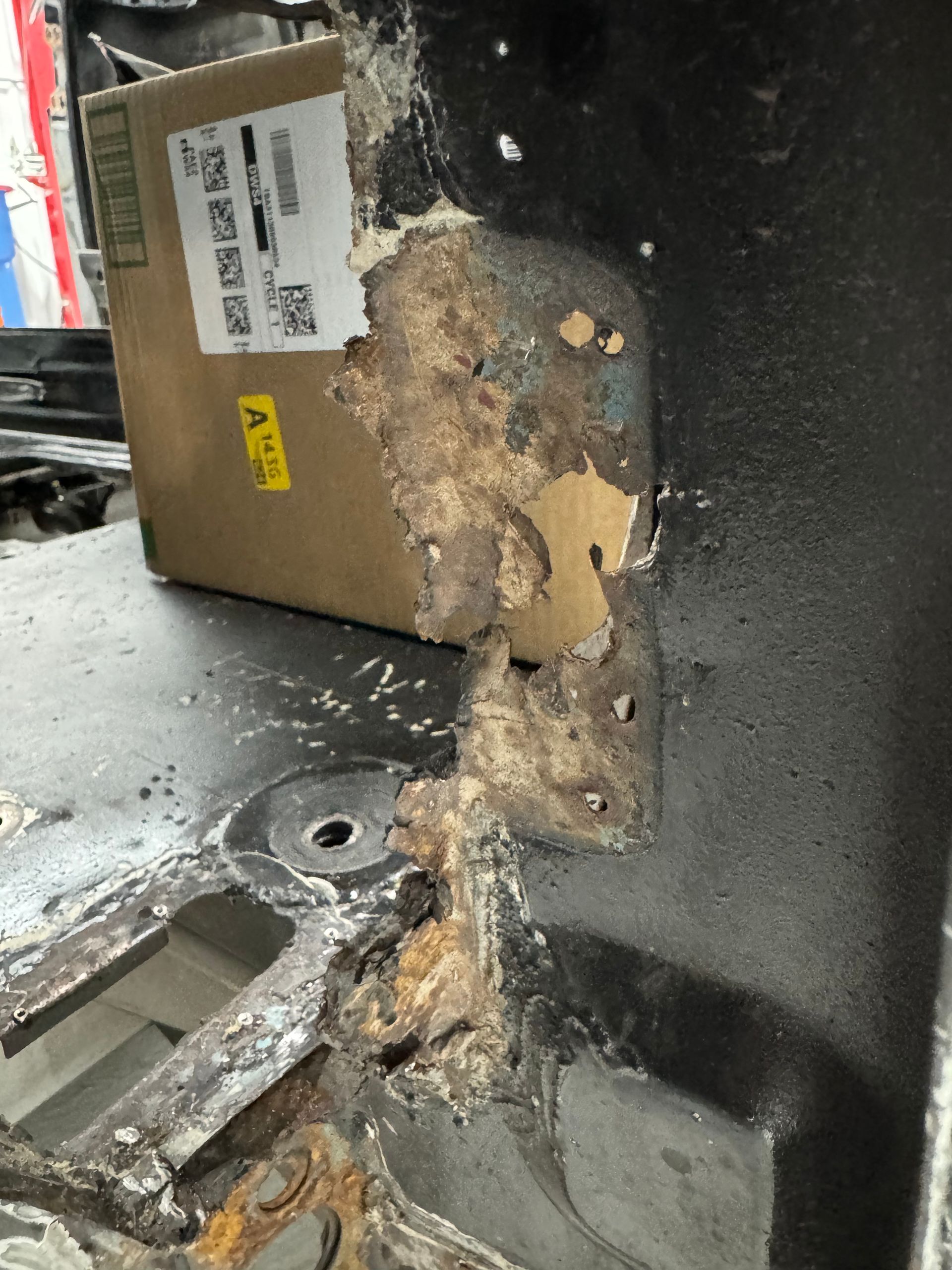 Rusty, corroded metal corner with peeling brown residue beside a black panel and cardboard box.