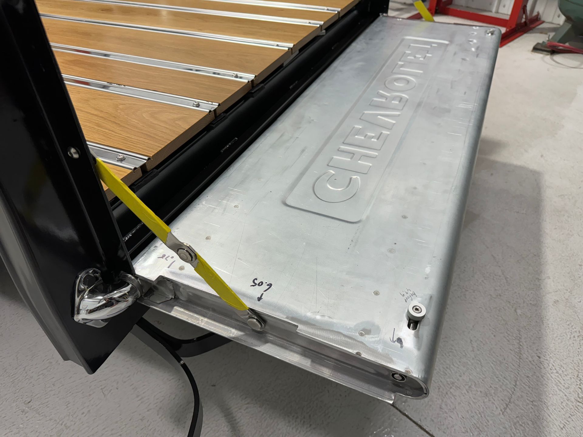 Silver forklift attachment labeled “GILLMANN” beside a black metal rack and wood slats in a workshop.