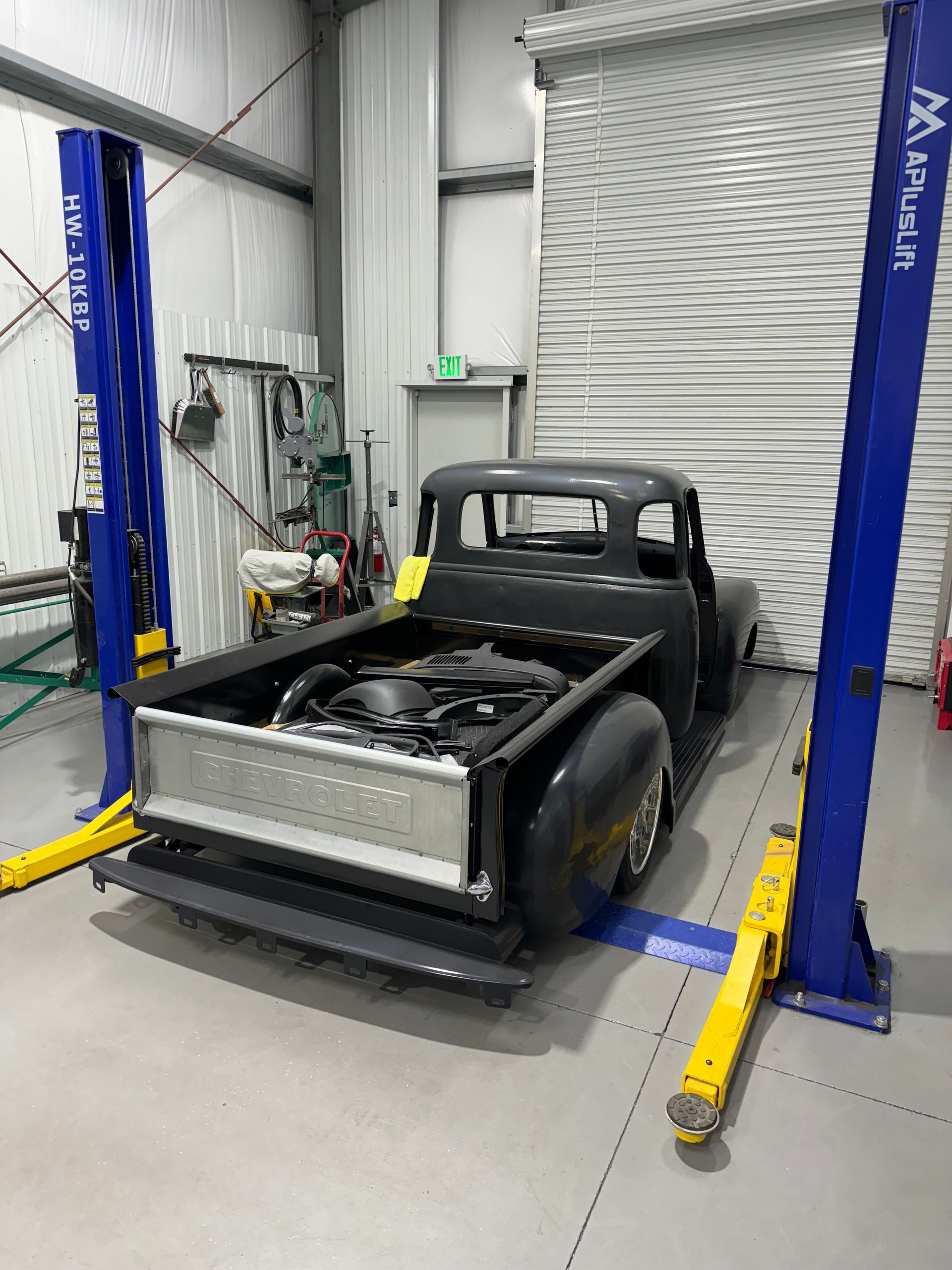 Black pickup truck in a garage lift bay with yellow arms and a closed roll-up door