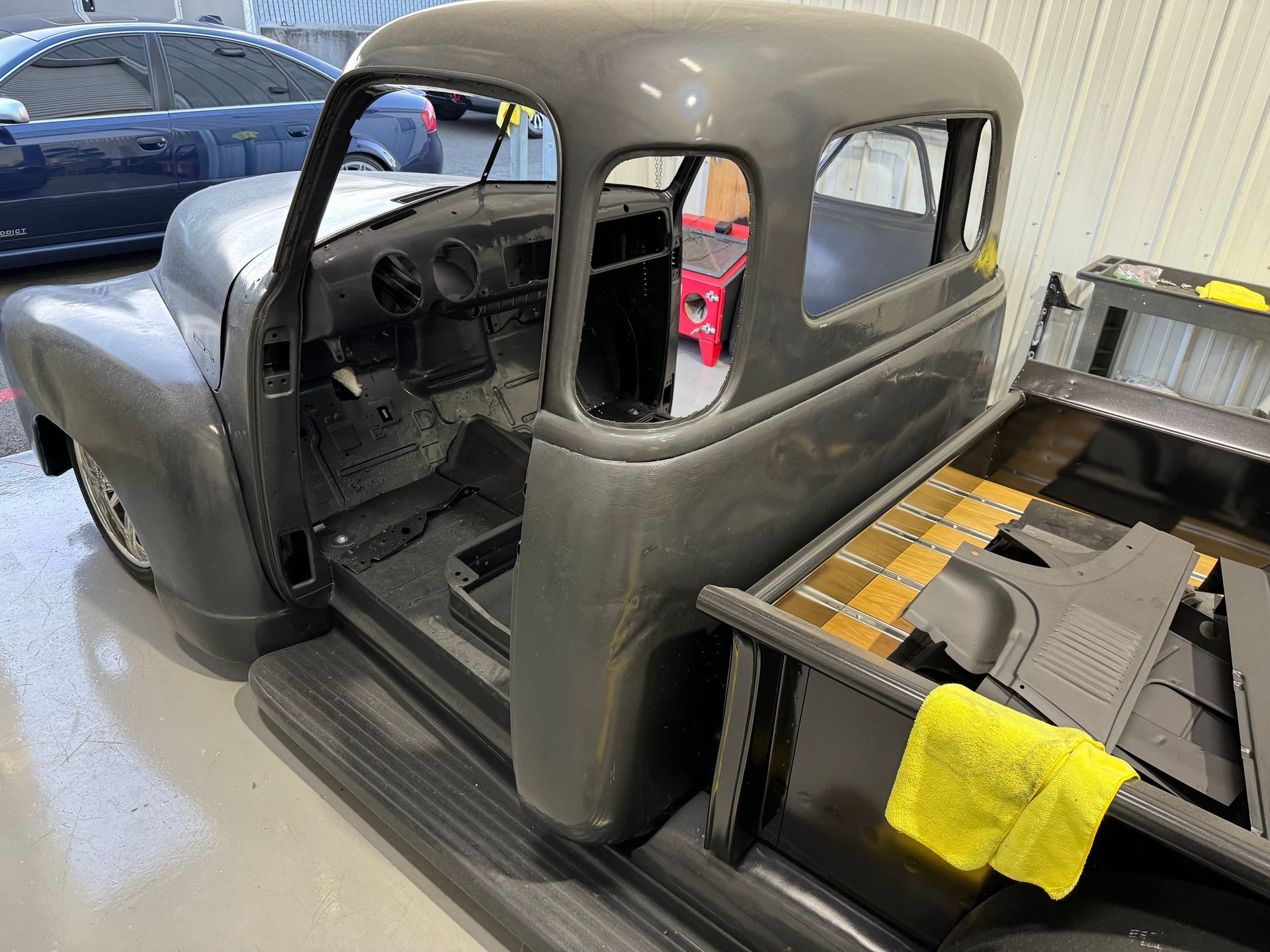 Gray vintage truck cab in a workshop next to a partially built wooden flatbed with a yellow rag
