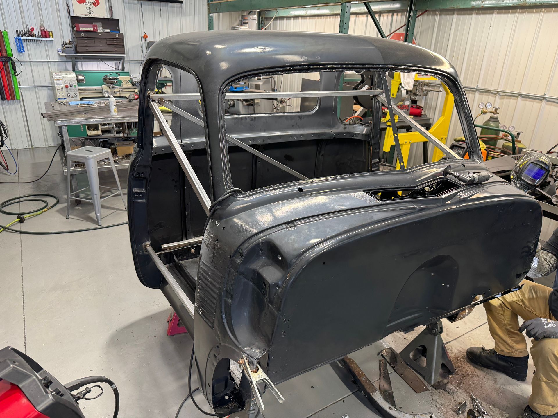Classic car body shell in a workshop, with a mechanic working beside it and tools in the background.