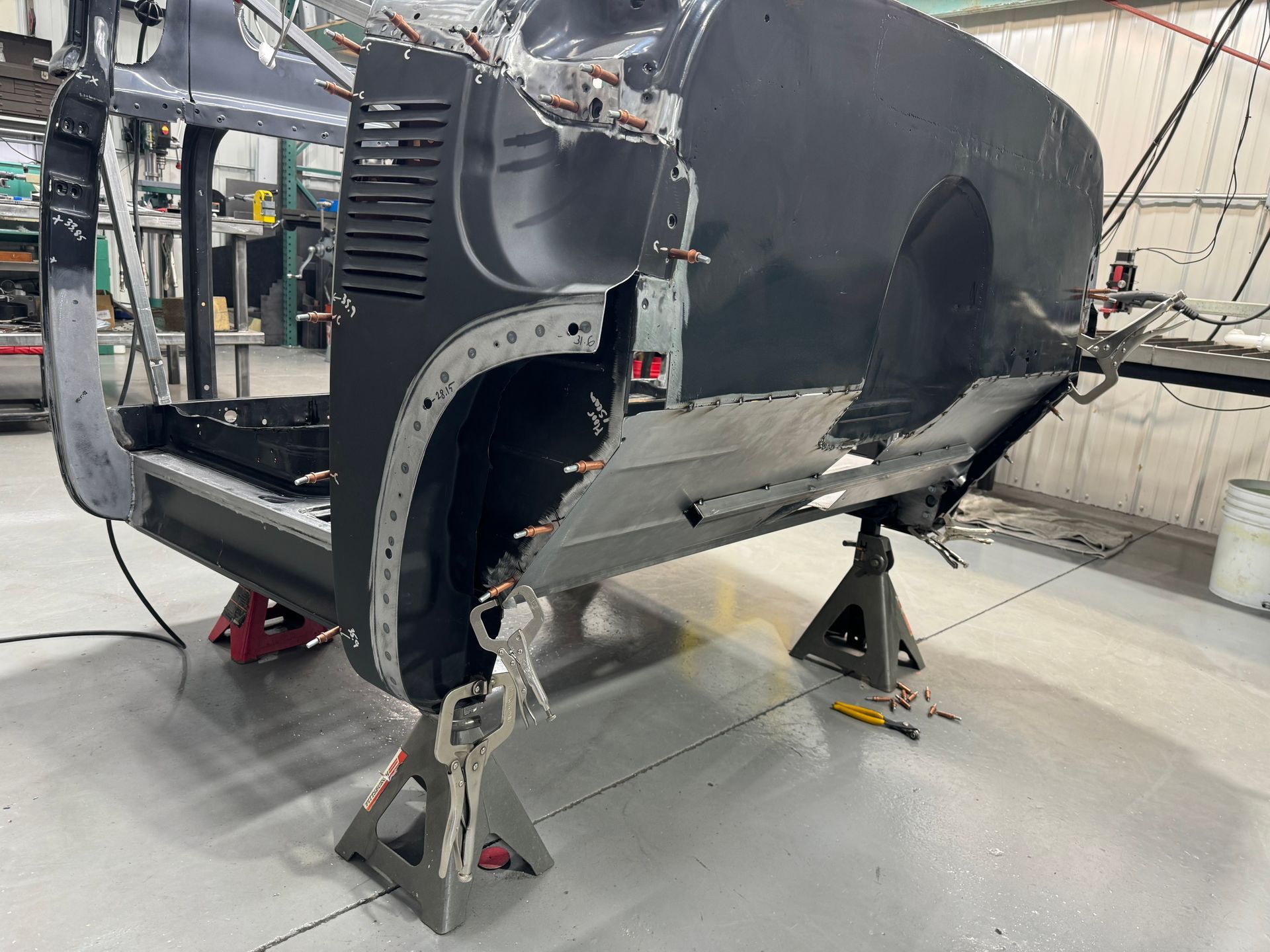 Partially assembled black vehicle body on stands in a workshop, with exposed frame and wheels removed