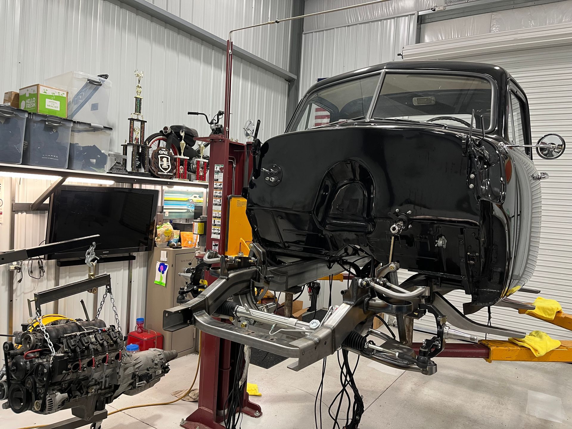 Classic car body on a lift in a workshop, with engine parts and tools scattered nearby.
