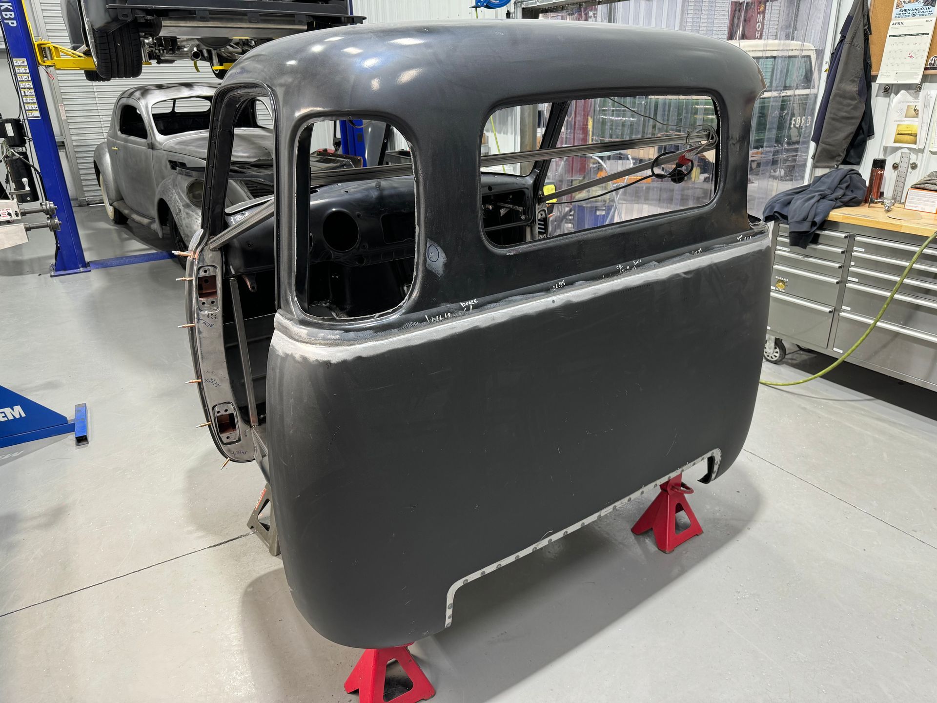 Gray vintage truck cab body on red jack stands in an auto workshop, with a white vehicle in the background