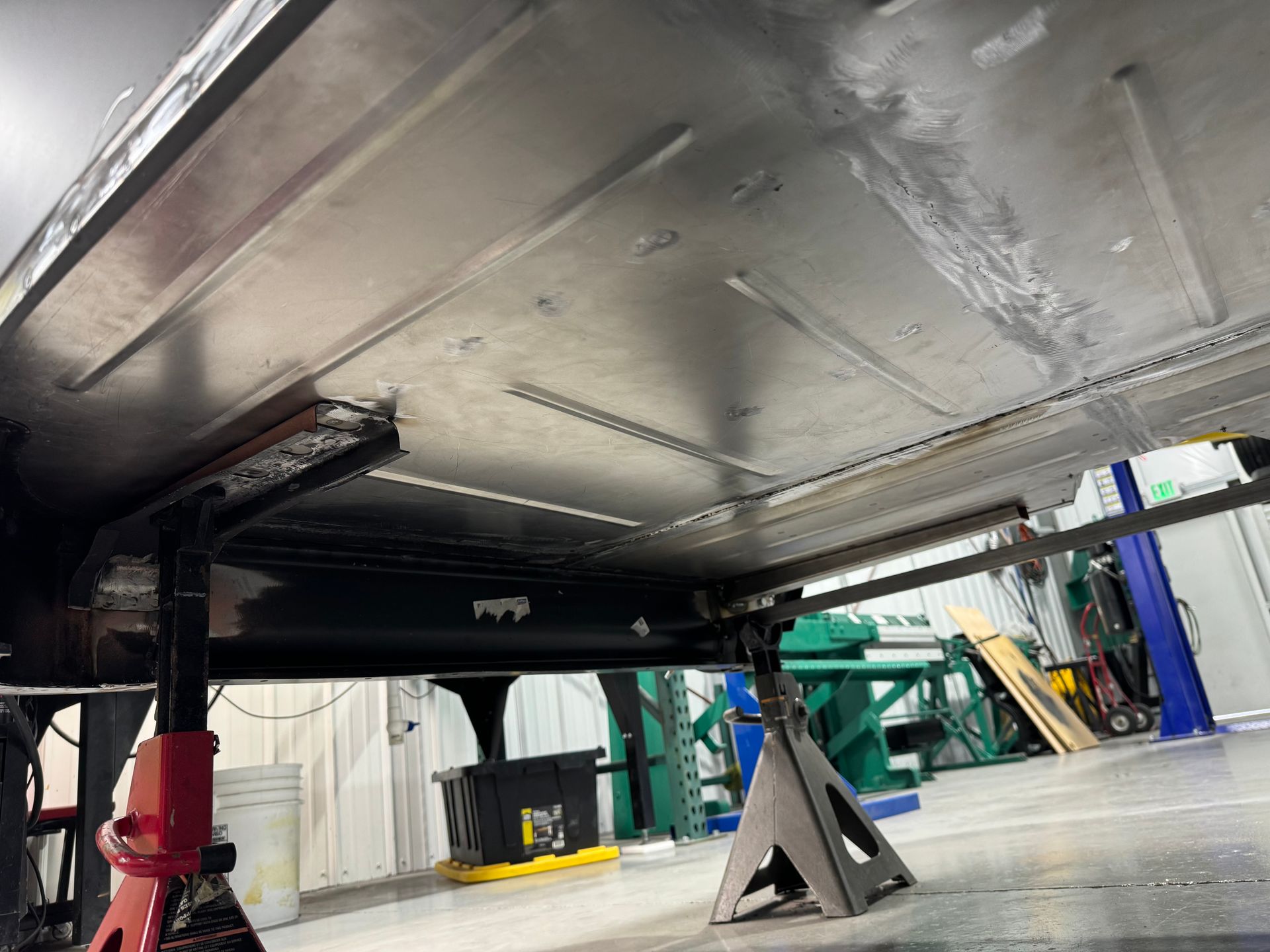 Underbody of a car on jack stands in a garage, with a lift and workshop equipment nearby.