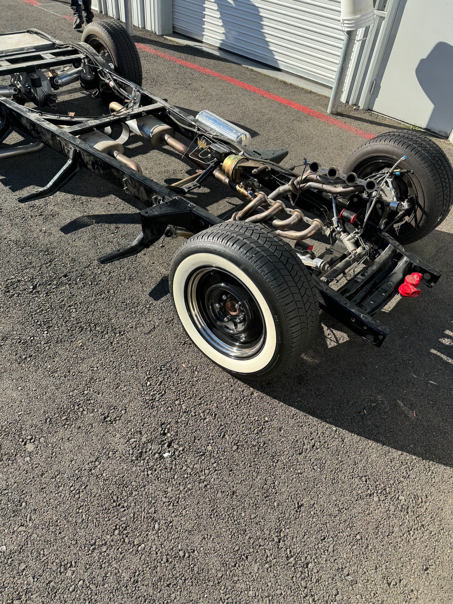Bare vehicle chassis with wheels on a gravel surface beside a building wall