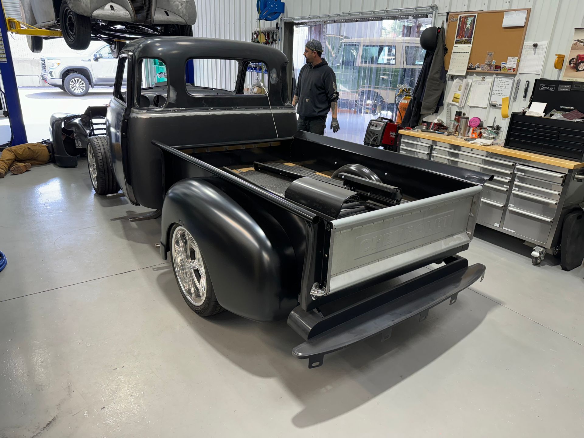 Black vintage pickup truck in a workshop, rear bed exposed with a person standing beside it