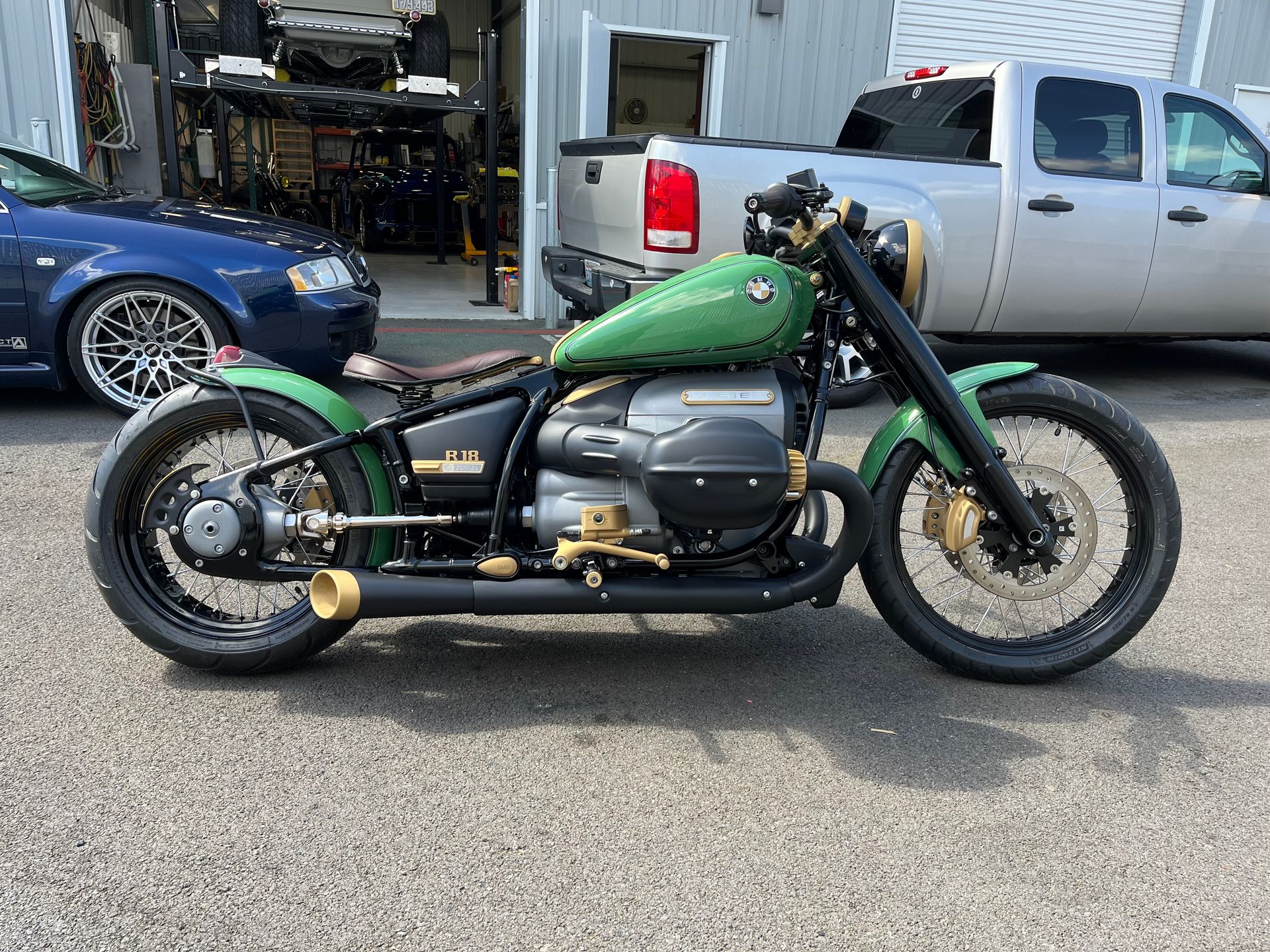 Green custom motorcycle parked outdoors in front of a garage, with black frame and gold exhaust.