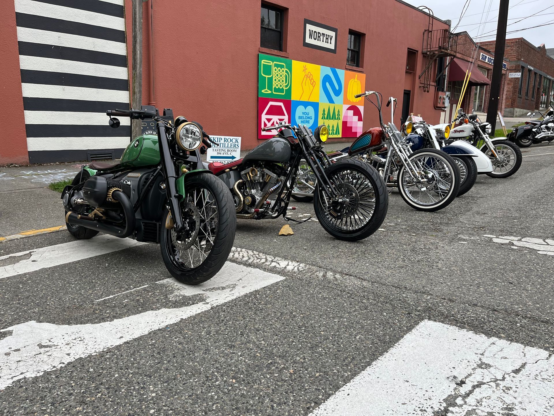 Row of parked motorcycles on a city street beside a brick wall with colorful mural panels.